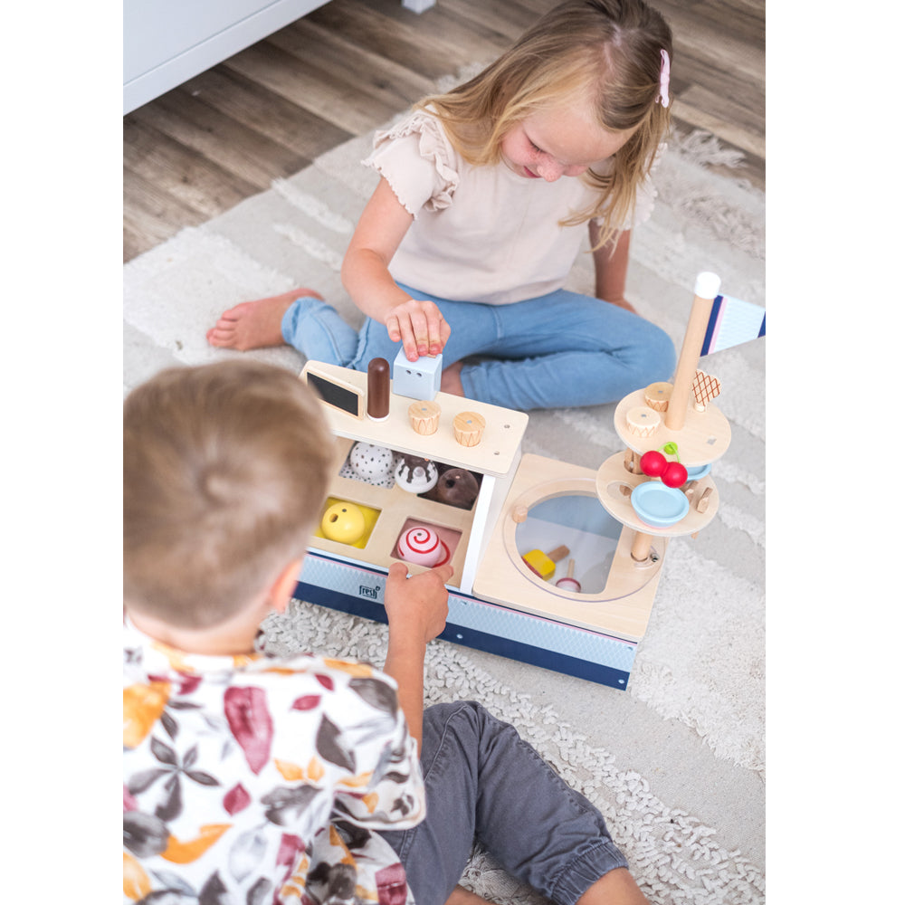 Zwei Kinder spielen mit der Eistheke „fresh“ Holzspielzeug-Eisdiele auf hellem Teppichboden.