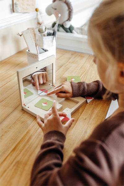Ein Kind interagiert mit dem Symmetriespiel mit Spiegel „Educate“ auf einem Holztisch, indem es mit seinen Fingern Muster anordnet, die im Spiegel reflektiert werden.