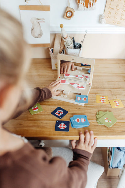 Ein Kind spielt das Symmetriespiel mit Spiegel „Educate“, ein Kartenspiel mit einem Spiegel auf einem Holzschreibtisch, während Tierkunst die Wand schmückt.