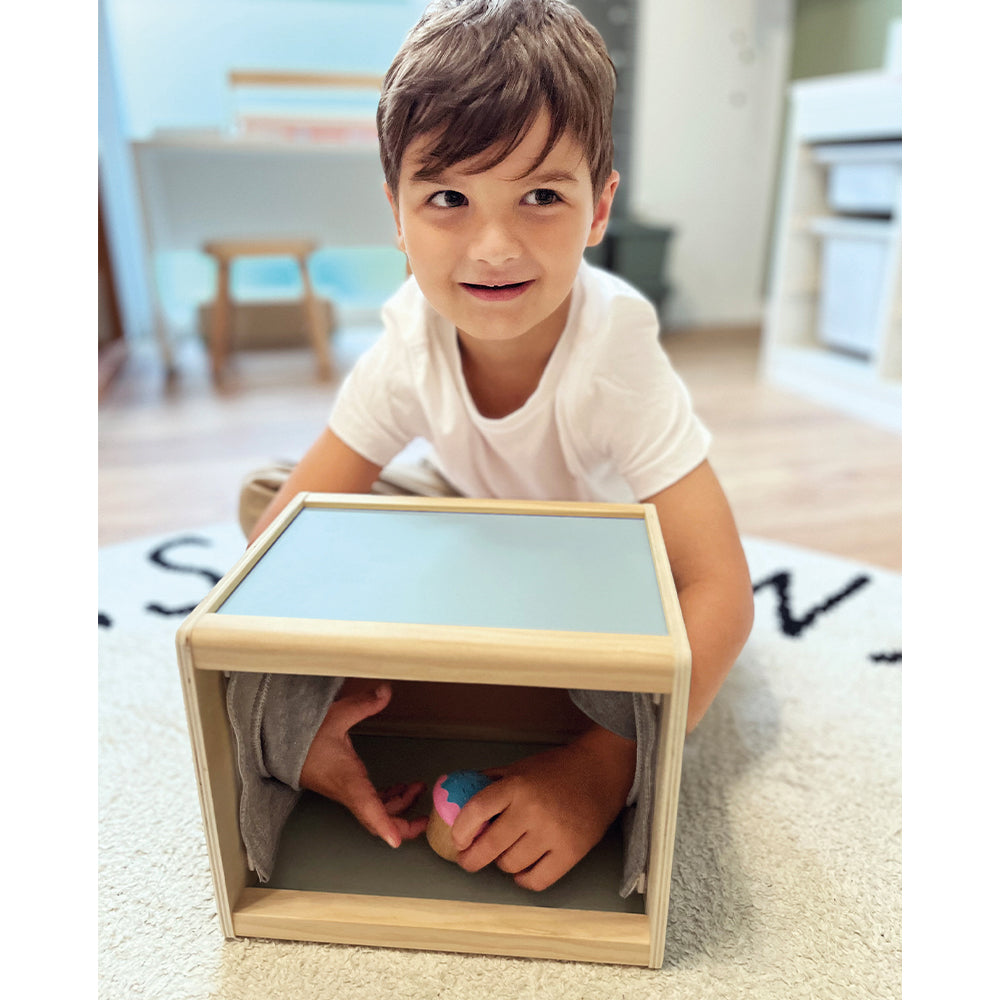 Ein lächelnder Junge kniet auf einem Teppich und greift in die Fühlbox „Sensory“, um nach einem darin versteckten Gegenstand zu tasten.