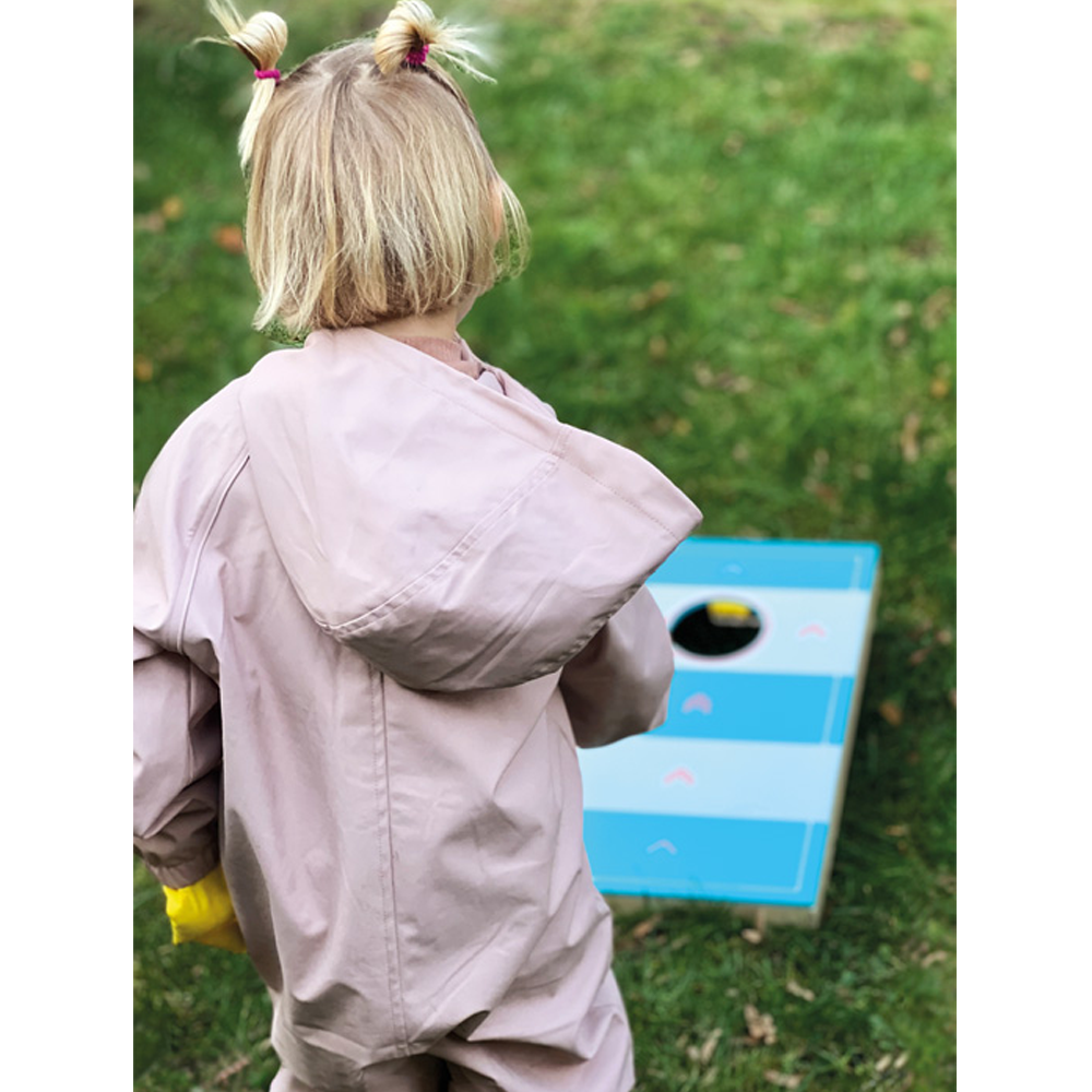 Ein Kind in einem rosa Regenmantel steht auf dem Gras und spielt mit dem blau-weißen Spielbrett „Active“ des Cornhole und Sling Puck Spiels.