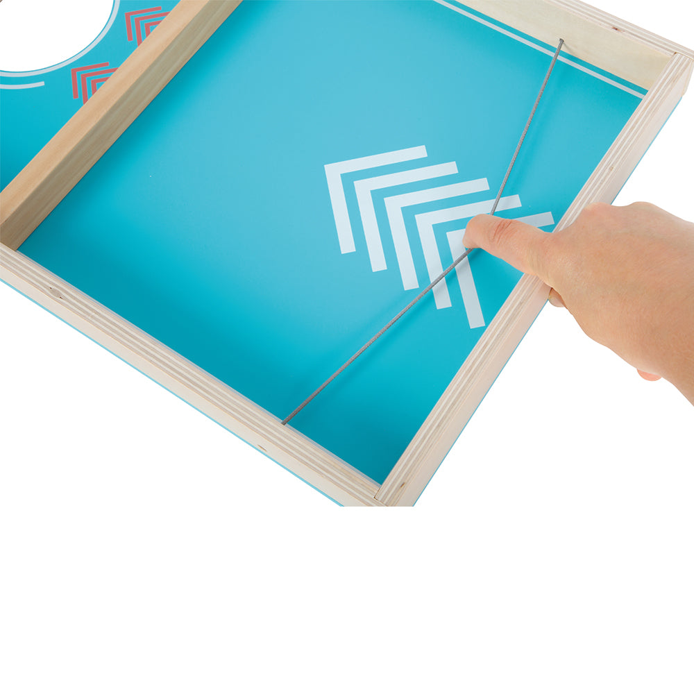 Eine Hand zieht ein Gummiband am Cornhole und Sling Puck Spiel „Active“ zurück, einem blauen Holztischspiel mit weißen geometrischen Mustern.