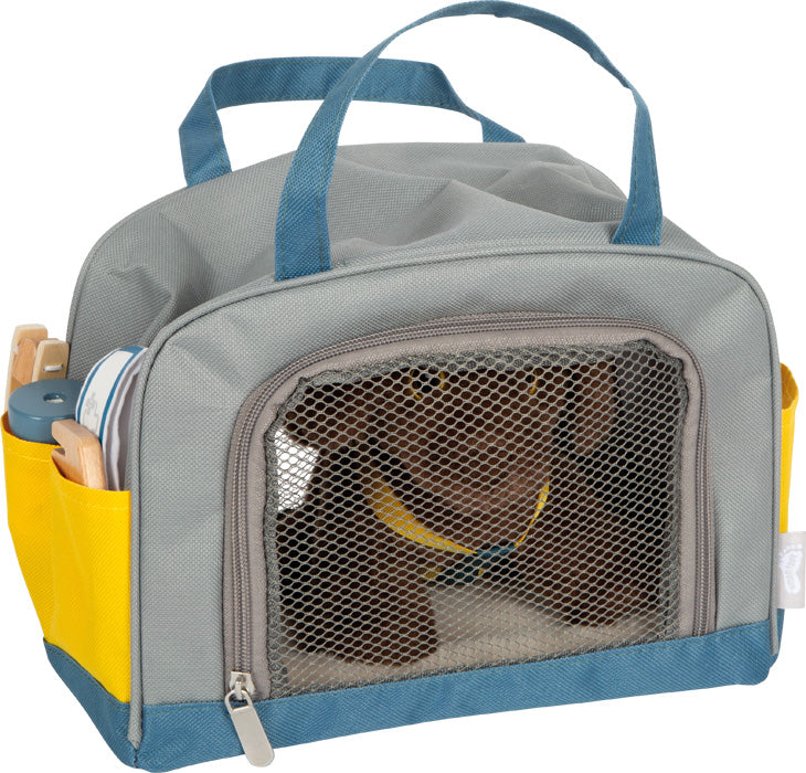 In einer grauen Tiertragetasche mit gelben Seitentaschen sitzt ein kleiner brauner Stoffhund aus dem „Hund mit Tasche und Pflege-Set“.
