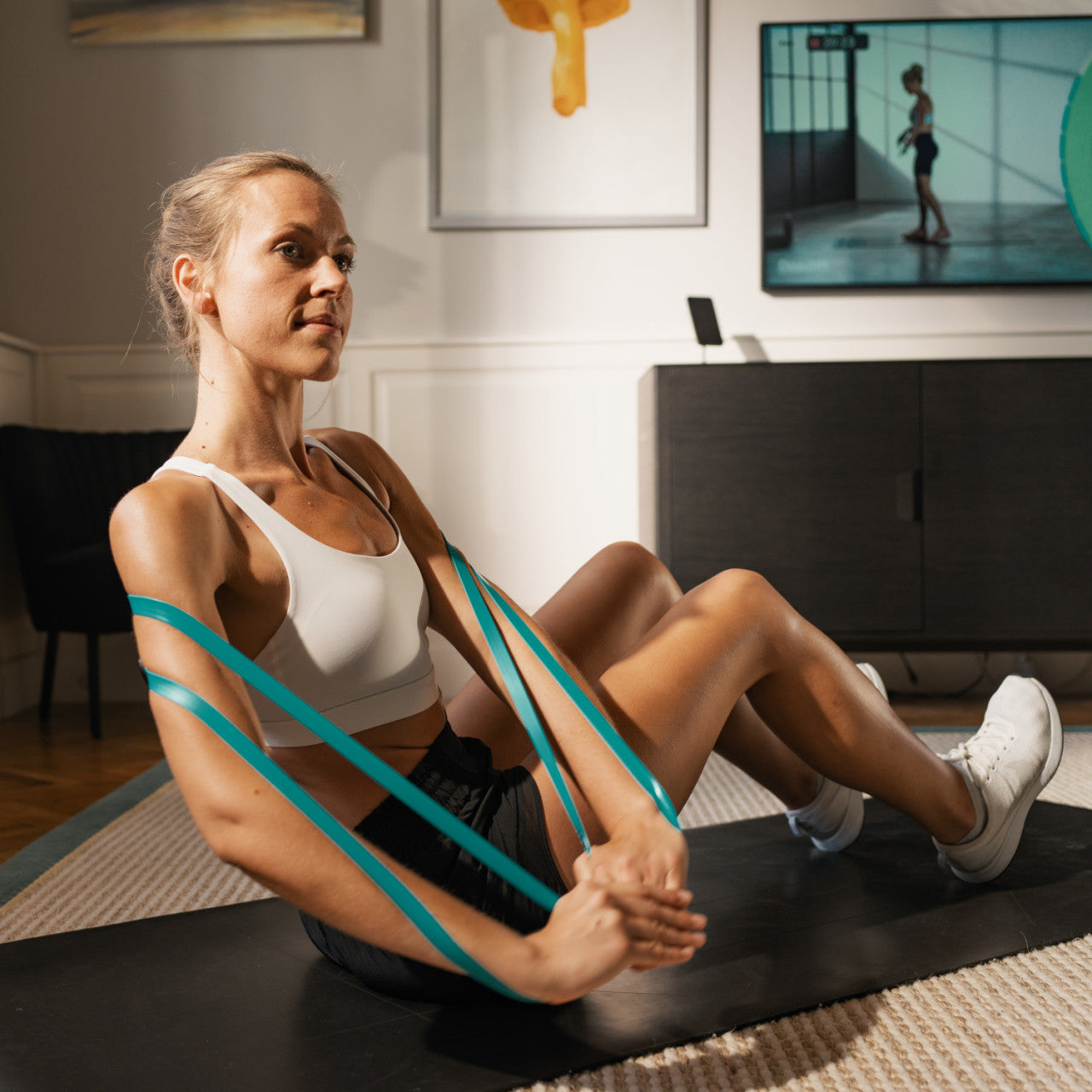 Frau trainiert zu Hause mit dem Smartes Fitnessband STRAFFR mit App - mittel, sitzt auf einer Matte und verfolgt ein Fitnessvideo im Fernsehen.
