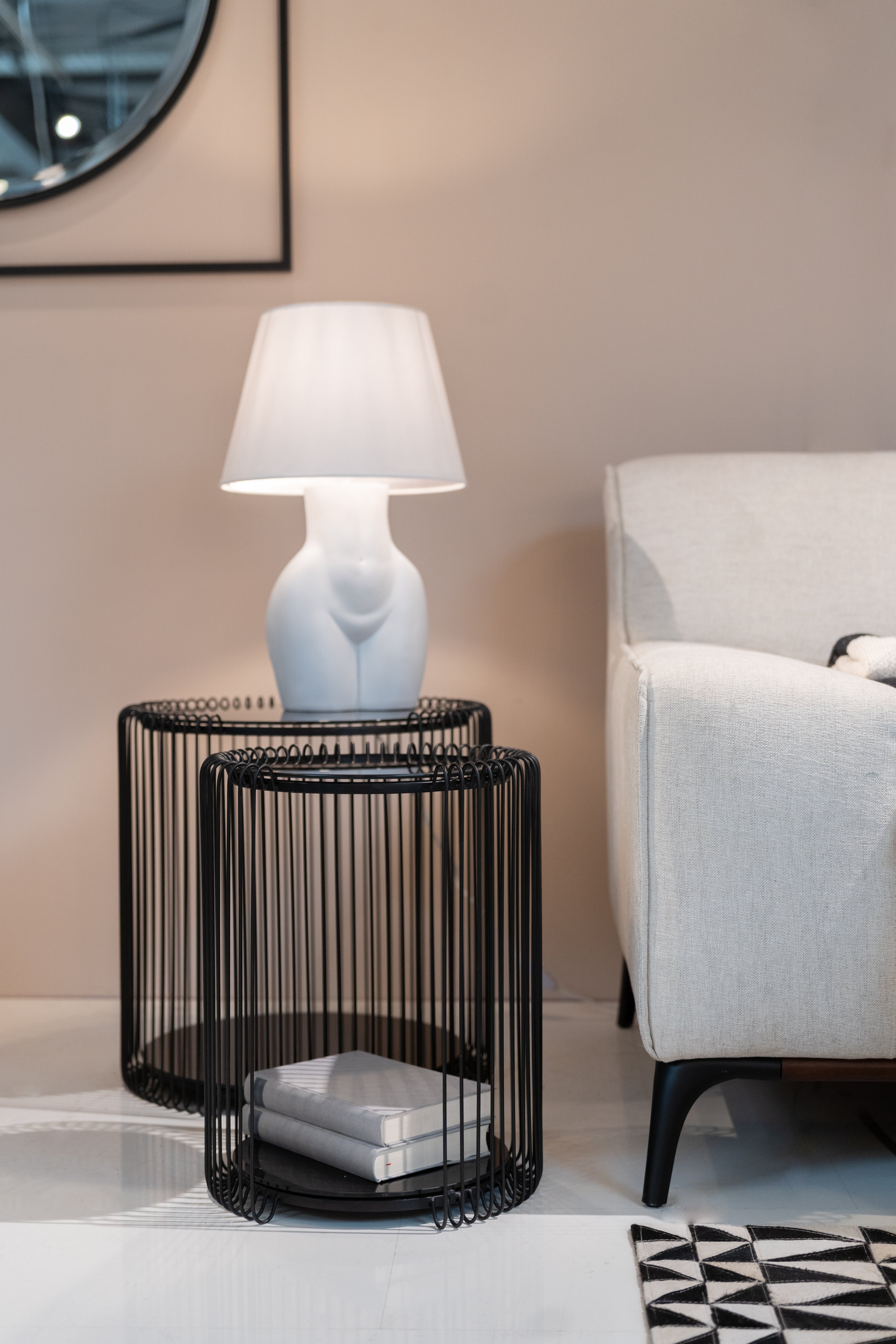 Ein modernes Wohnzimmer mit dem Beistelltisch Wire Double Schwarz (2/Set), einer weißen skulpturalen Lampe, zwei Büchern, einem hellbeigen Sofa und einem Teil eines geometrischen Teppichs auf einem glänzenden Boden – perfekt für modernes Design.