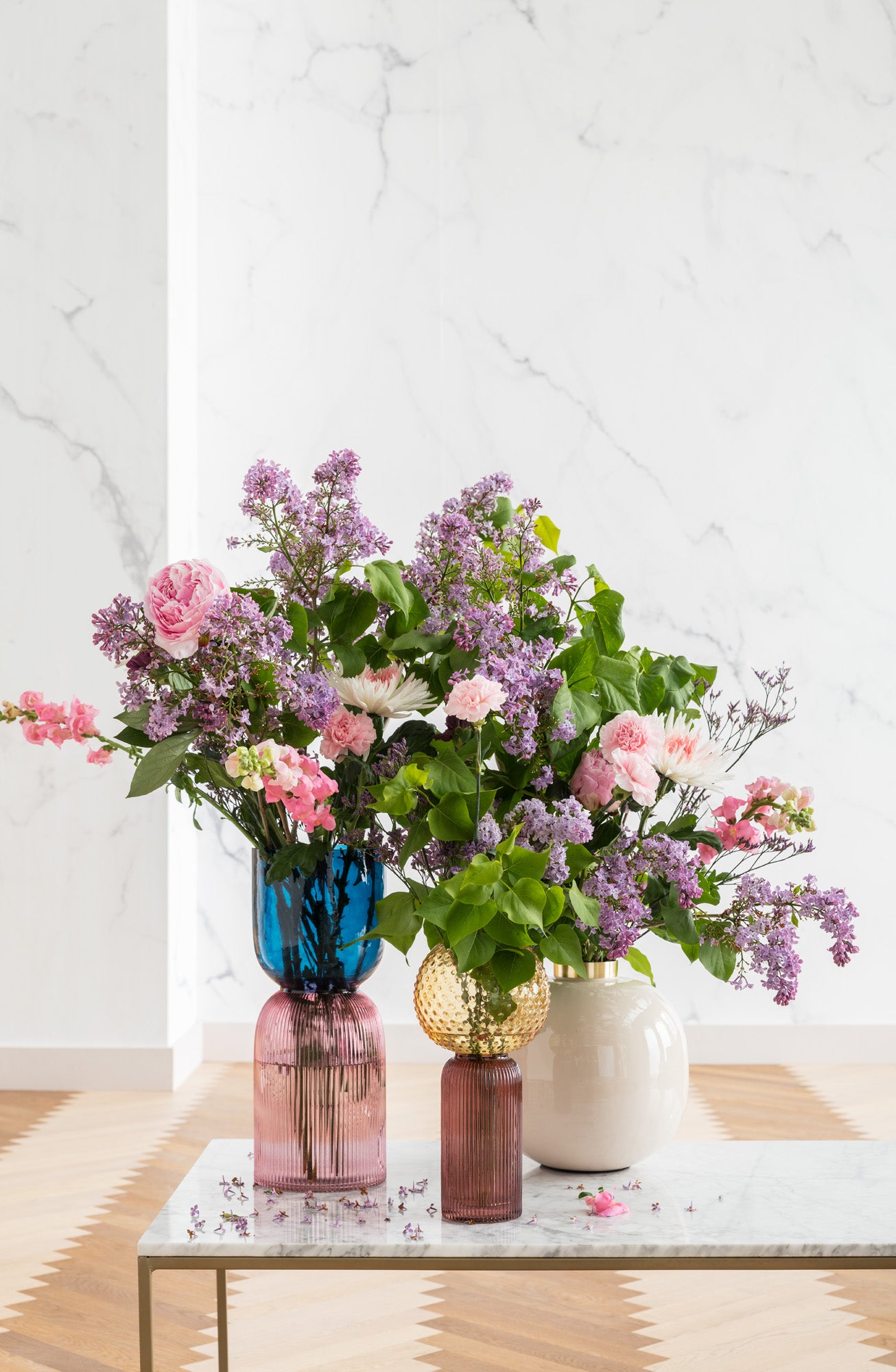 Drei Vasen „Marvelous Duo Blau Lila“, jede mundgeblasen und mit rosa und violetten Blumen gefüllt, sind auf einem Tisch mit Marmorplatte in einem hellen Raum mit einer hellen Wand mit Marmormuster und einem Holzboden im Chevron-Muster arrangiert.