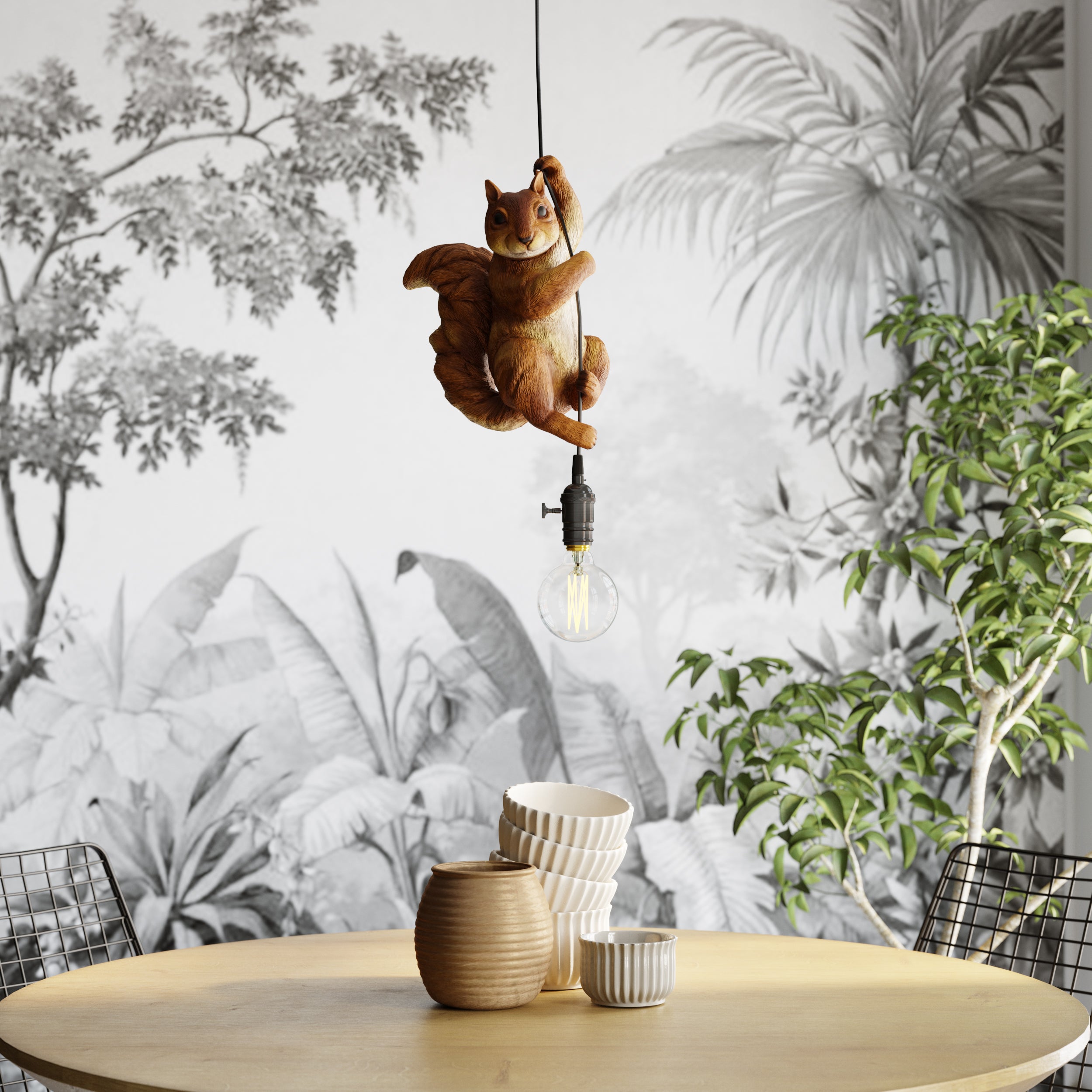 Eine skurrile Hängeleuchte „Animal Squirrel“ hängt über einem runden Holztisch mit Keramikschüsseln und -tassen, vor dem Hintergrund einer schwarz-weißen tropischen Tapete und einer grünen Blattpflanze.