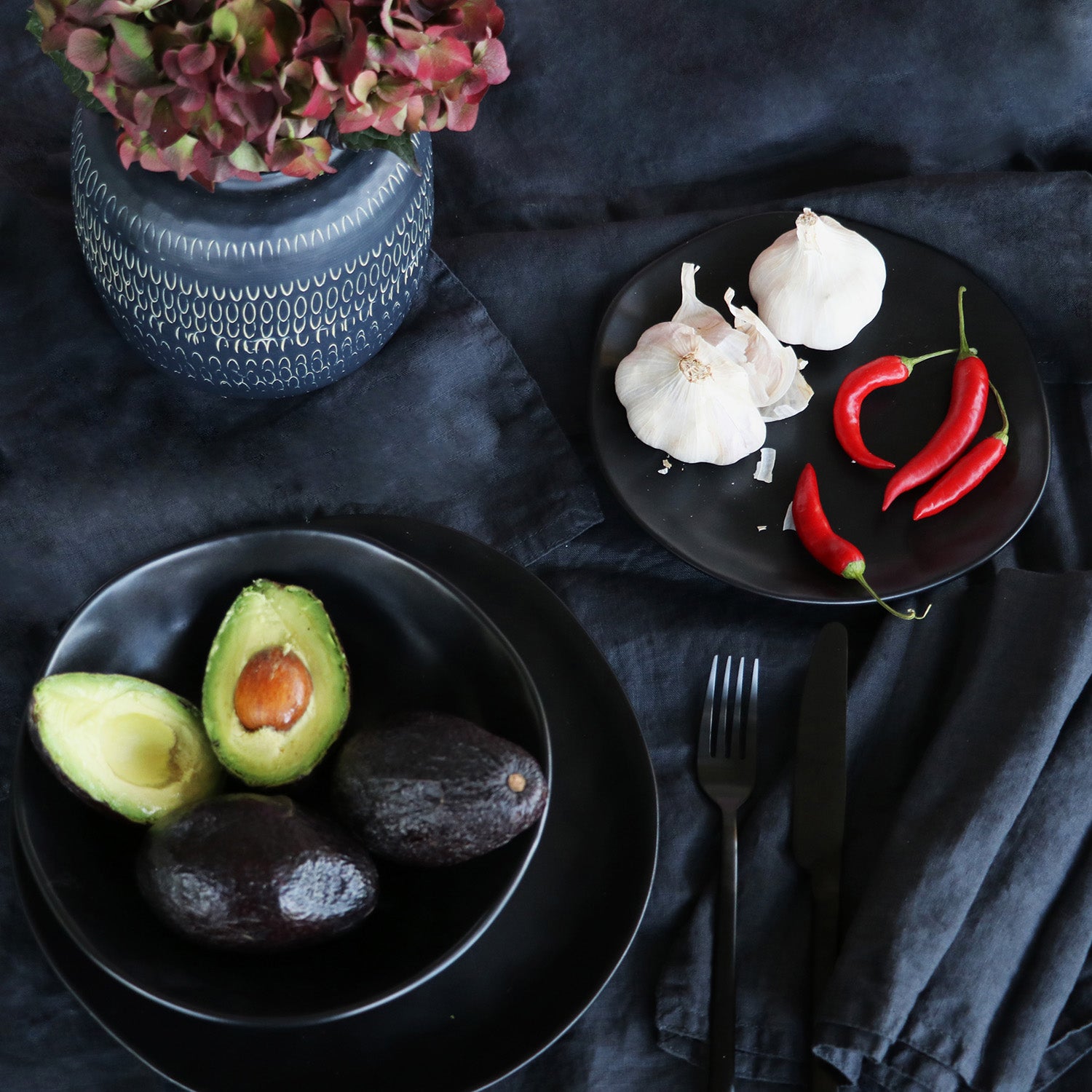 Ein Tischgedeck in dunklem Design punktet mit dem Teller Organic Black Ø20cm in Hand-made-Optik, einer Vase mit Hortensien, Knoblauchknollen und roten Chilischoten auf einem Teller, drei Avocados – eine davon aufgeschnitten – auf einem anderen sowie einer eleganten schwarzen Gabel und einem Messer.