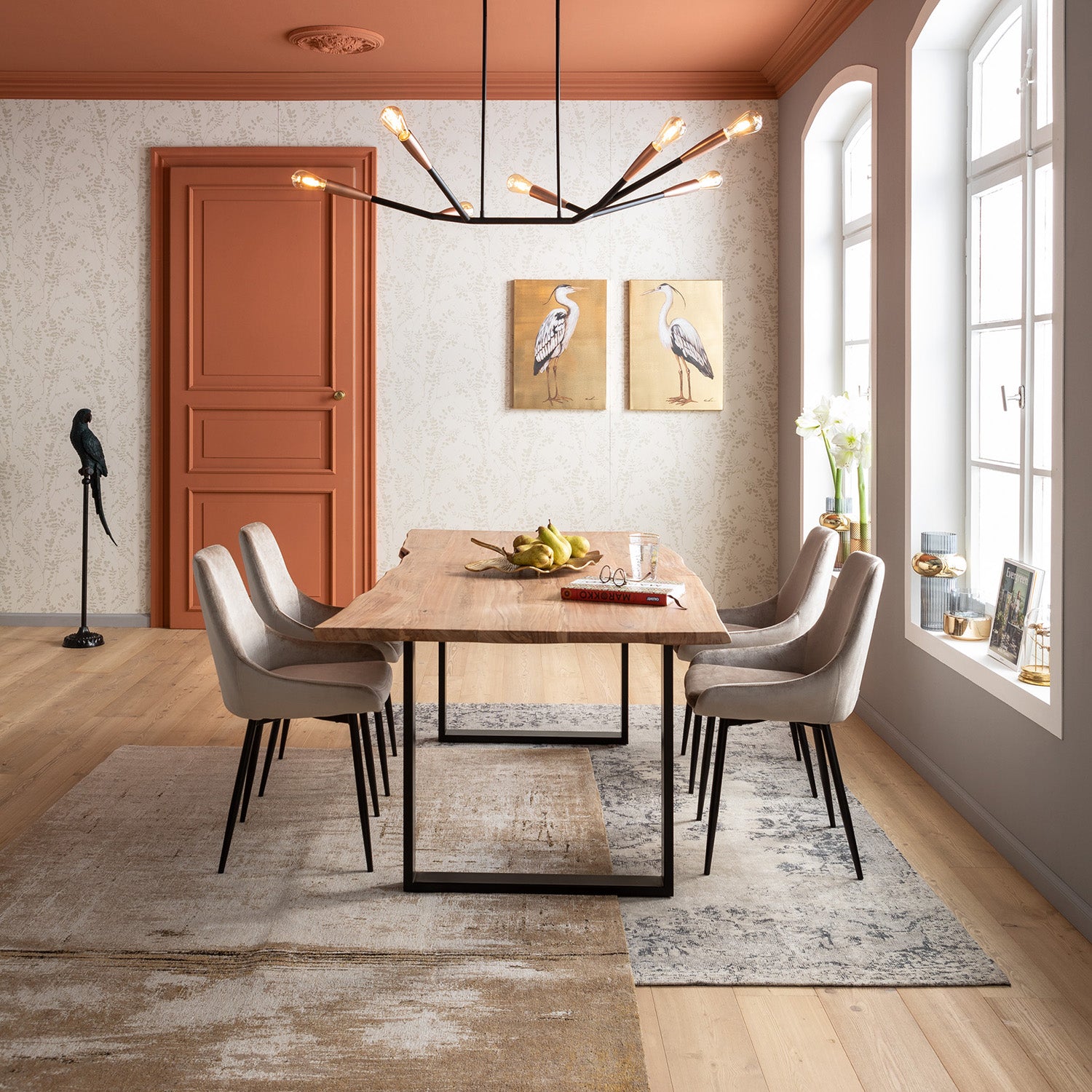 Modernes Esszimmer mit hellem Holztisch, sechs Stuhl East Side Pearl Samtstühlen und einem schwarzen Kronleuchter. Große Fenster lassen Sonnenlicht herein. Goldene Vogelkunst, eine Vogelstatue und eine Obstschale schaffen eine elegante Atmosphäre.