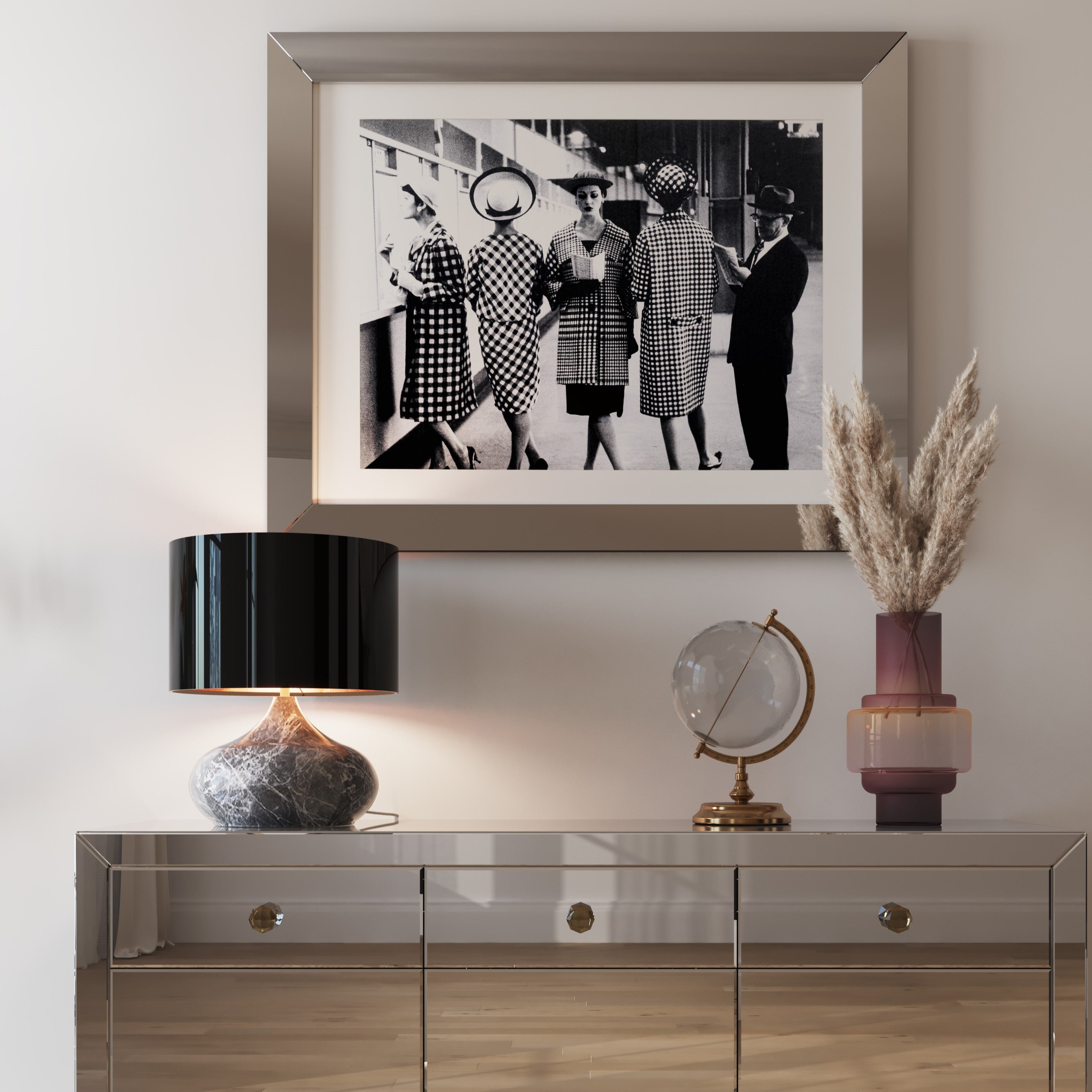 Ein verspiegelter Konsolentisch mit einer Tischleuchte Mamo Deluxe, einem Deko-Globus und Pampasgras in einer Vase. Darüber hängt ein Schwarz-Weiß-Foto von Frauen in Vintage-Outfits an der beigefarbenen Wand.