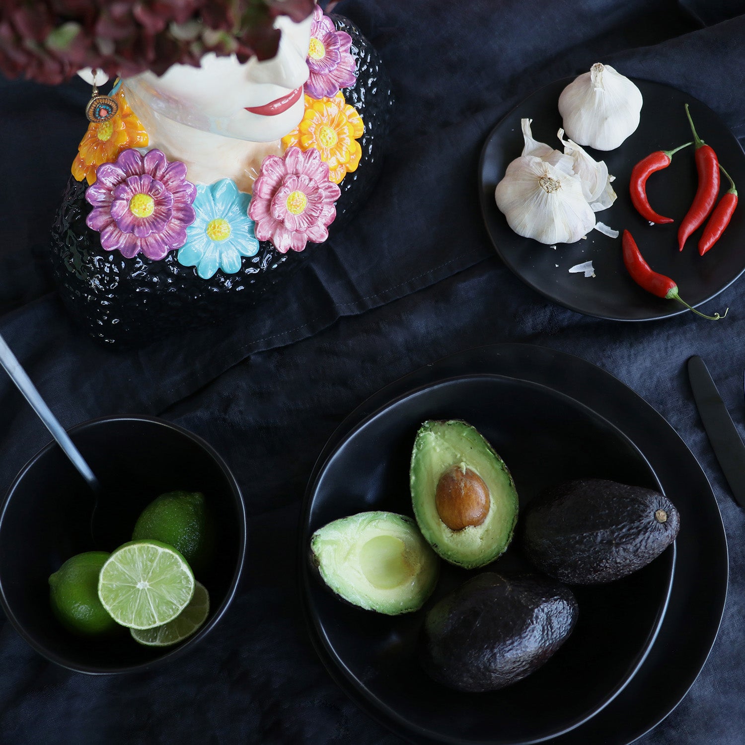 Eine dunkle Tischdekoration mit Keramikgeschirr: Eine Schale mit Limetten, eine Schale mit Avocados (eine aufgeschnitten), Knoblauchknollen und roten Chilischoten auf dem Teller Organic Black Ø26cm, dazu eine Blumenvase mit aufgemaltem Gesicht in der oberen linken Ecke.