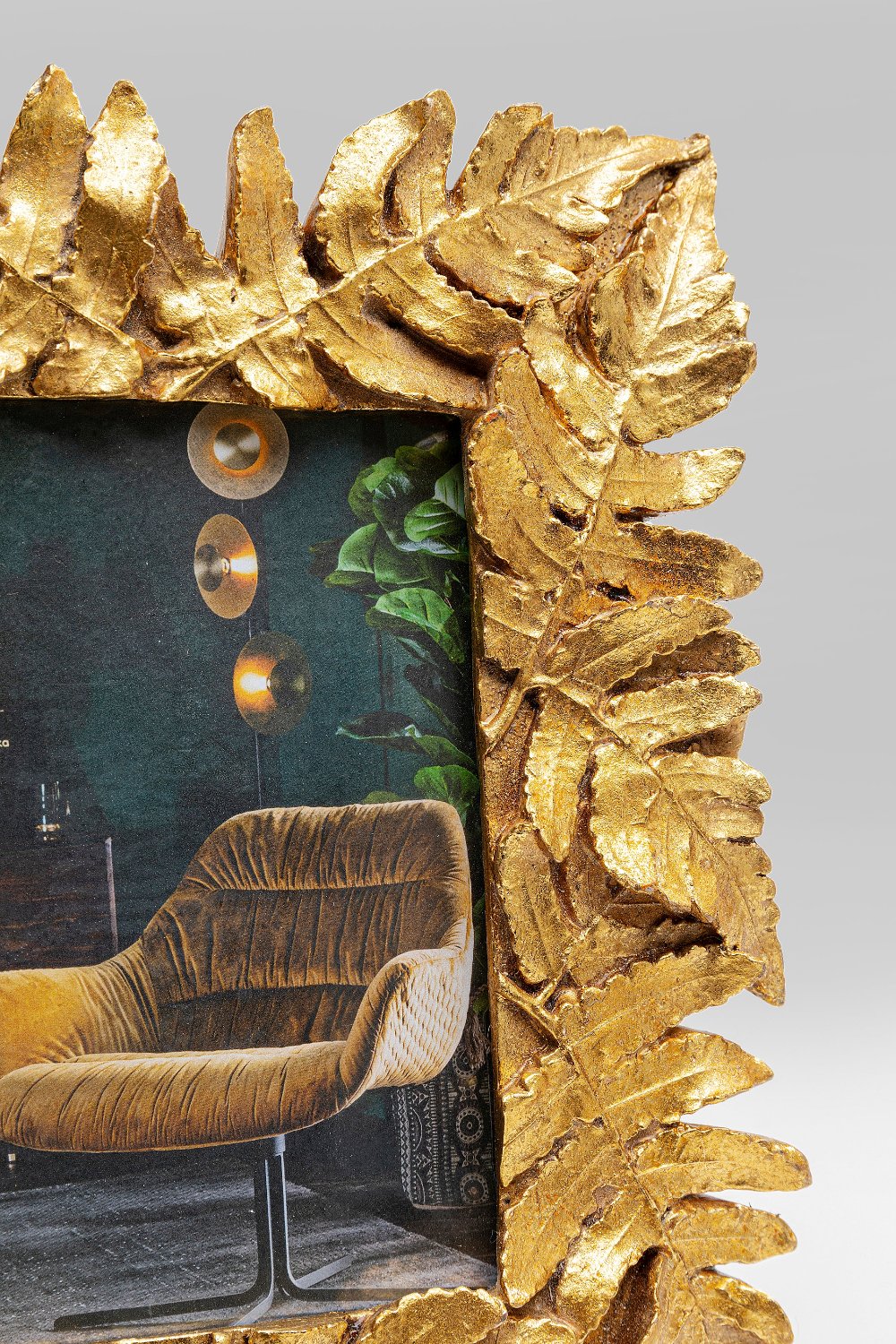 Nahaufnahme des Bilderrahmens Gold Leaves 10x15cm, handgefertigt aus Polyresin mit Goldfolie und Blattmotiven. Im Rahmen: ein Teil eines Samtsessels, eine dunkle Wand und Hängelampen.