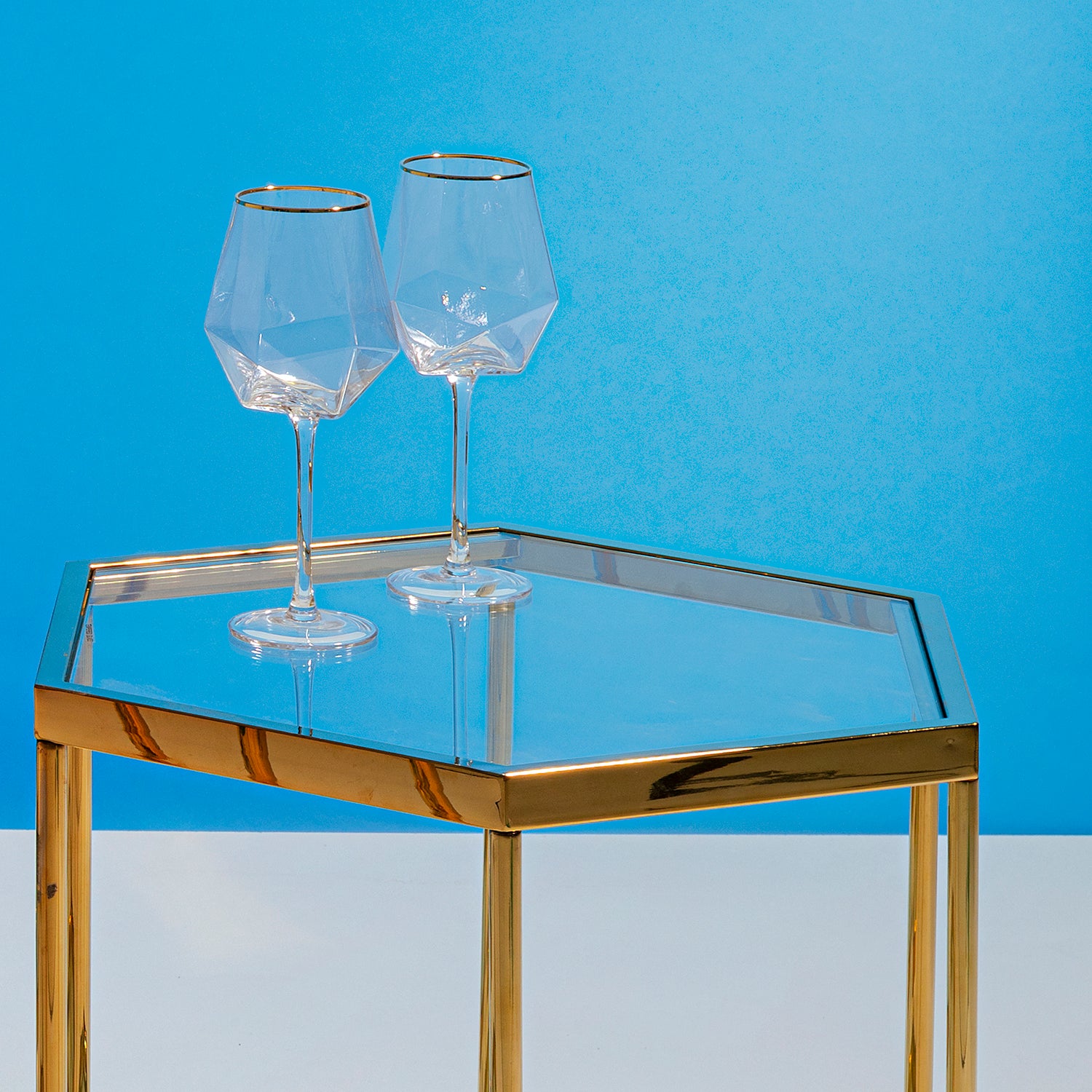 Zwei leere Weinglas Diamond Gold Rim (4/tlg.) stehen auf einem Tisch mit Glasplatte und Goldrahmen vor blauem Hintergrund und zeigen ihre elegante geometrische Form und den markanten Goldrand.