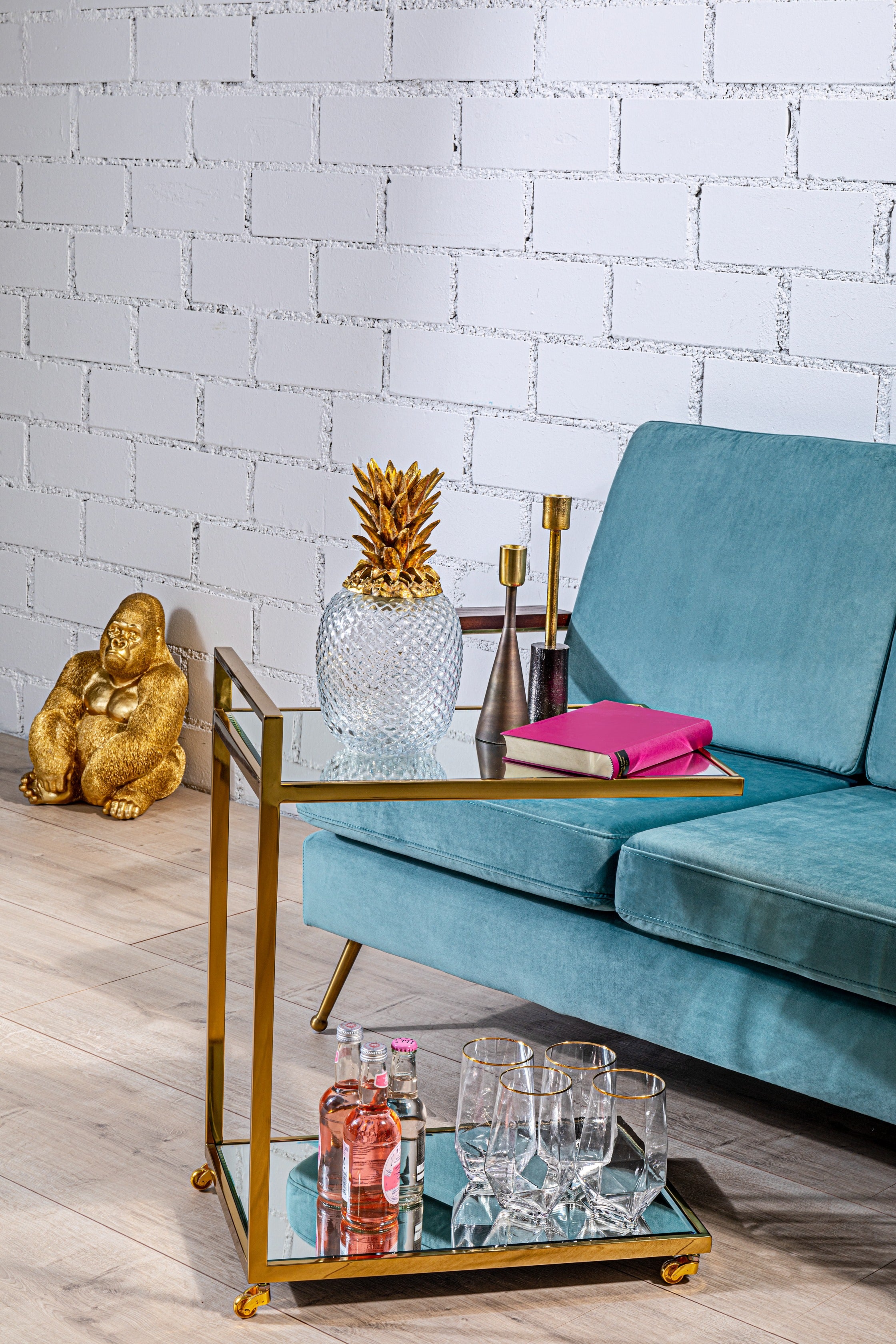 Ein modernes Wohnzimmer mit einem blaugrünen Sofa, einem Glas-Couchtisch mit einer Kristallananas-Dekoration, Büchern, Kerzen und einem 4-teiligen Wasserglas-Set mit Diamant-Goldrand. Auf dem Regal stehen Flaschen und Gläser; an der weißen Backsteinwand steht eine goldene Buddha-Statue.