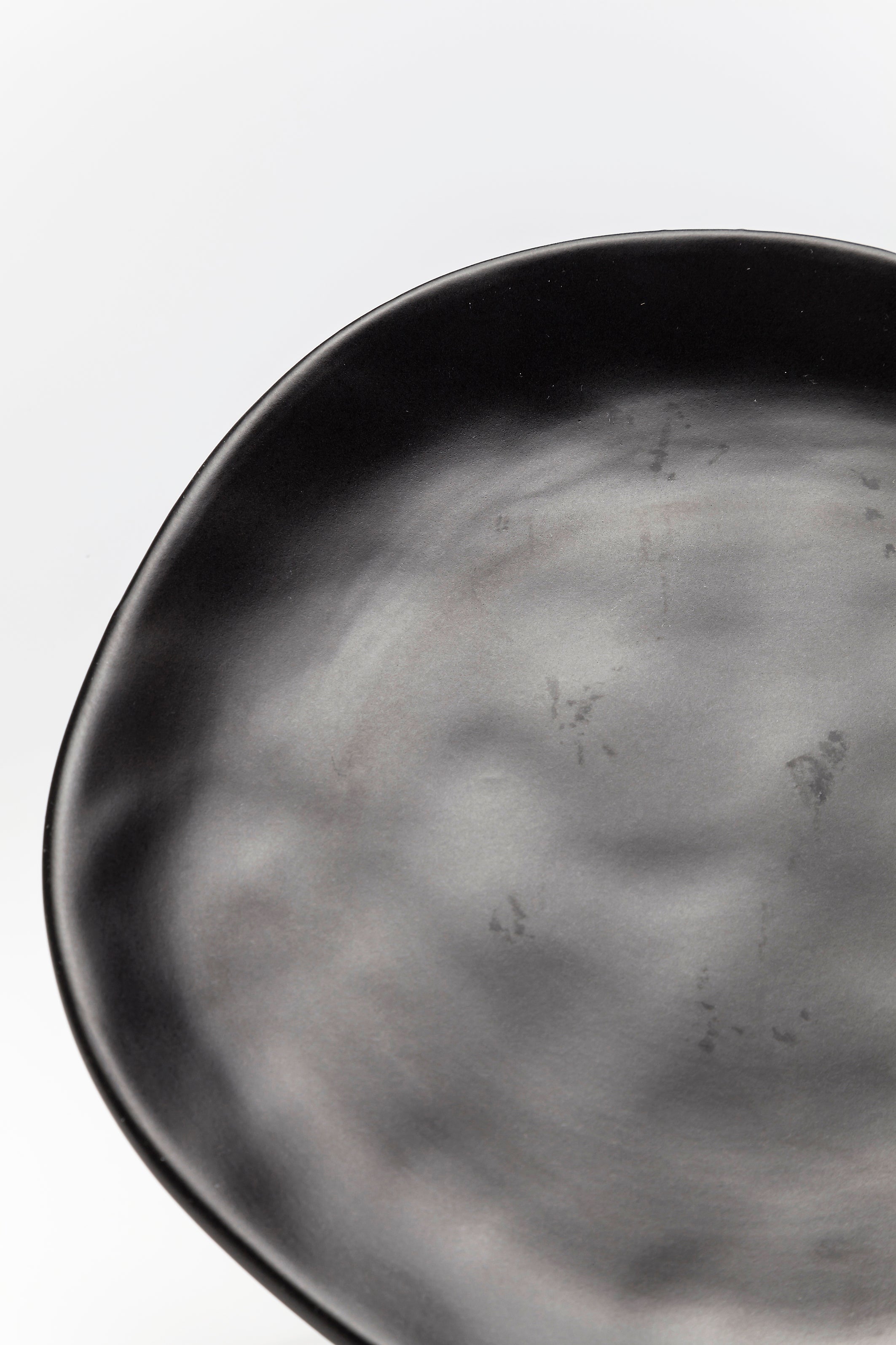 Nahaufnahme des Tellers Organic Black Ø20cm, ein mattschwarzer Steingutteller mit unregelmäßiger Form, der vor einem hellgrauen Hintergrund präsentiert wird, um seine subtile Oberflächenstruktur und die handgefertigten Optikreflexionen hervorzuheben.