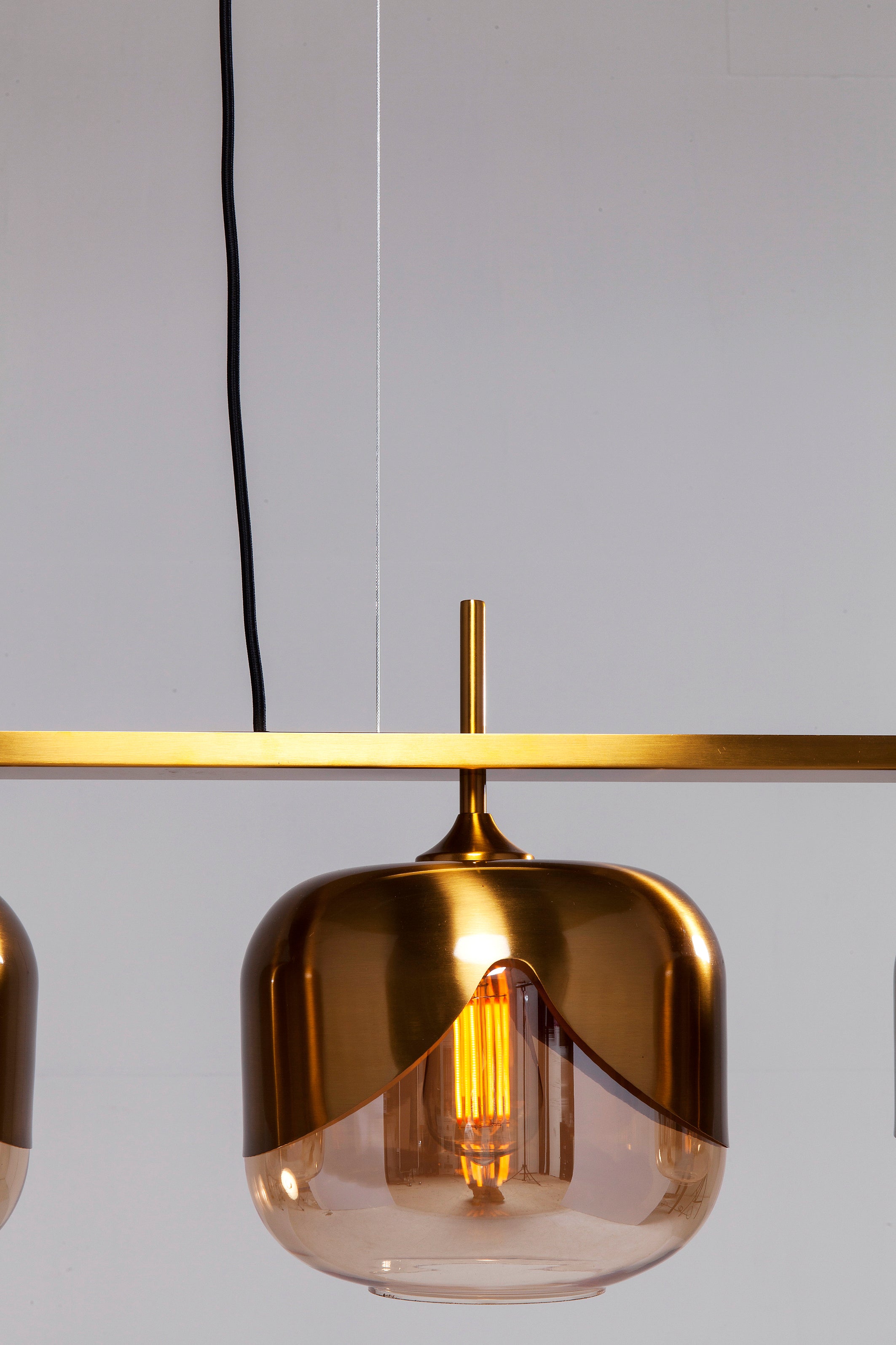 Die Hängeleuchte Golden Goblet Quattro Ø25cm verfügt über ein modernes Messing-Finish und einen klaren Glasschirm, der an einem schwarzen Kabel aufgehängt wird – ideal als elegante Esstischbeleuchtung mit ihrer sichtbaren, leuchtenden Glühbirne vor jeder hellen Wand.