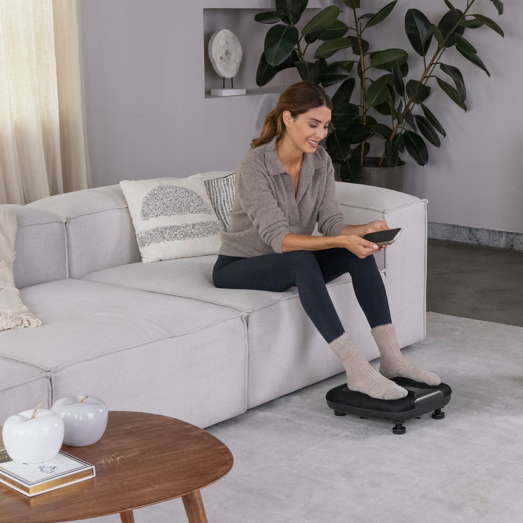 Frau sitzt auf einem Sofa, telefoniert und hat ihre Füße auf einer kleinen Fußstütze in einem gemütlichen Wohnzimmer.