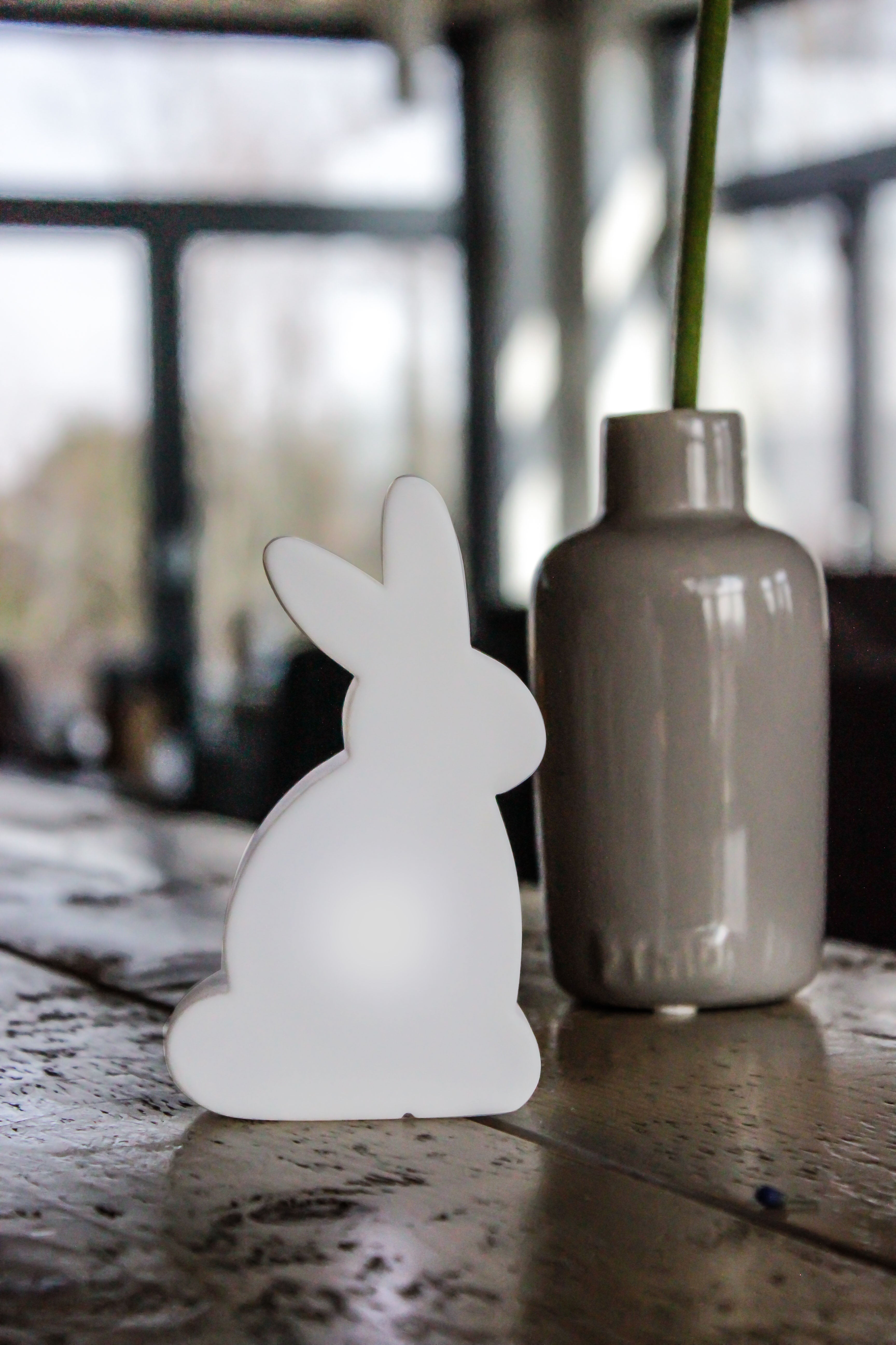 Weißes, kaninchenförmiges Objekt und eine graue Vase auf einer Holzoberfläche mit unscharfem Hintergrund.