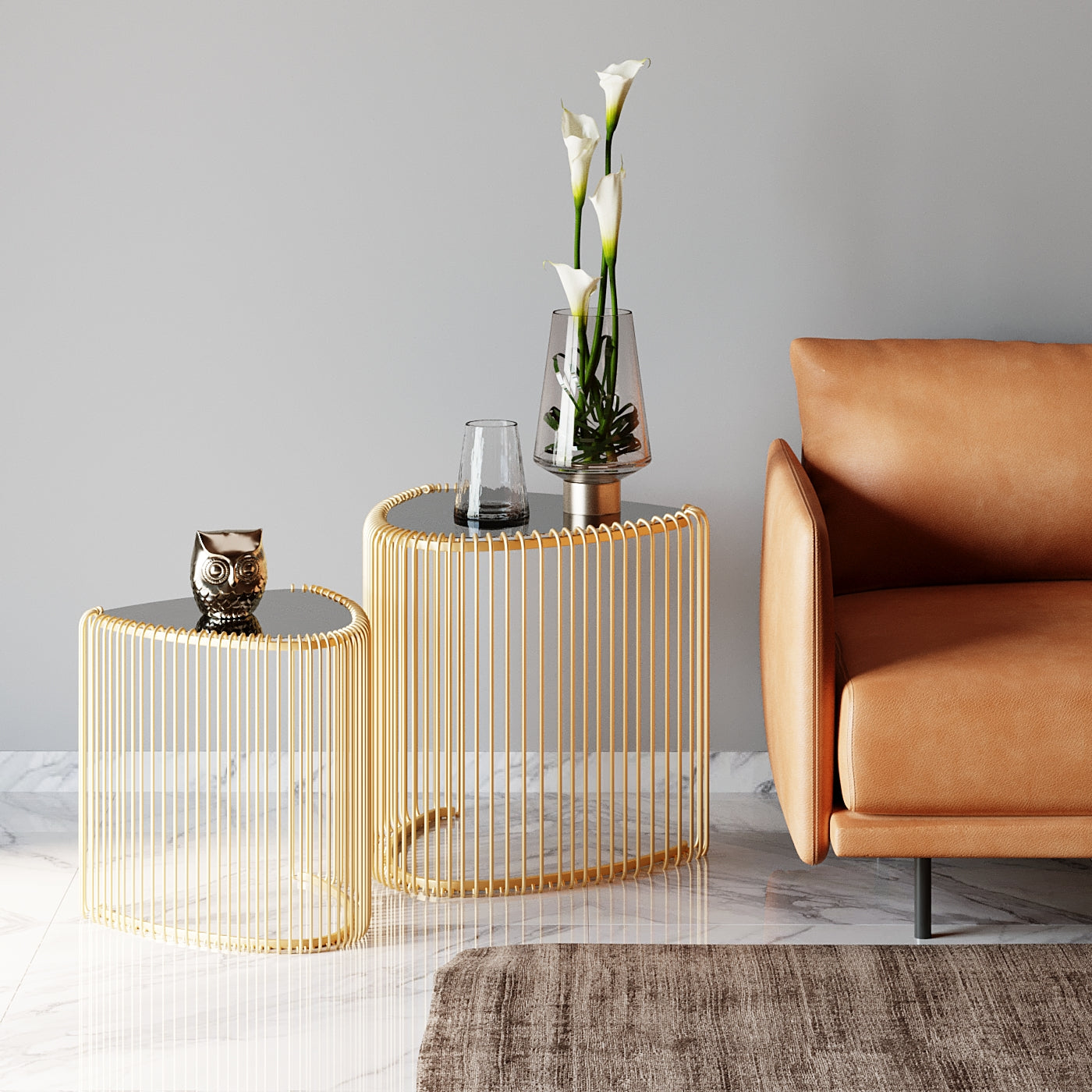Zwei moderne Beistelltische Wire Triangle Gold (2er-Set) mit Glasplatten und minimalistischem Design stehen neben einem hellbraunen Ledersofa. Einer präsentiert zwei Glasvasen mit weißen Callas, der andere zwei Kerzen. Marmorboden und ein beiger Teppich runden das Ambiente ab.