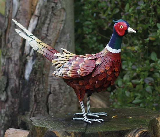 Die Gartenfigur „Fasan“ ist eine farbenfrohe Fasanenskulptur aus Metall, die auf einem Baumstumpf mit viel Grün im Hintergrund steht.