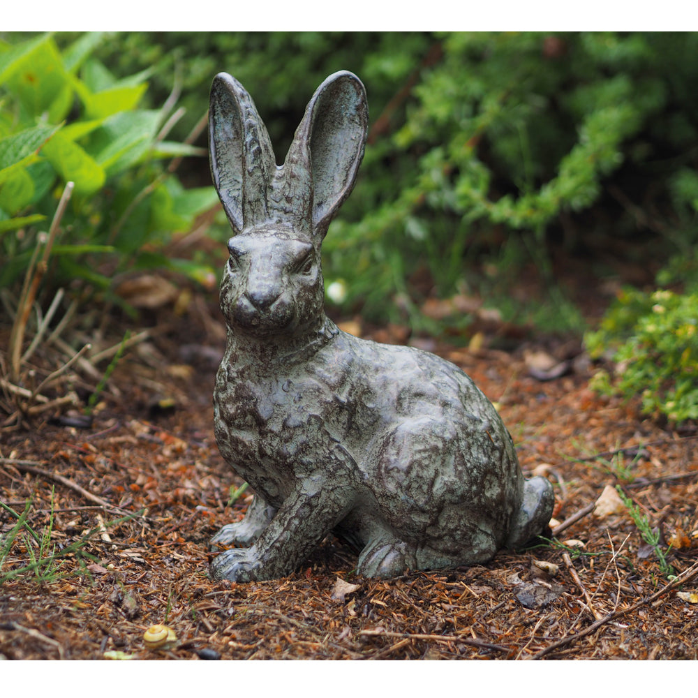 Die Gartenskulptur „Hase“ aus Bronze steht im Garten auf dem Boden und ist von grünen Pflanzen umgeben.