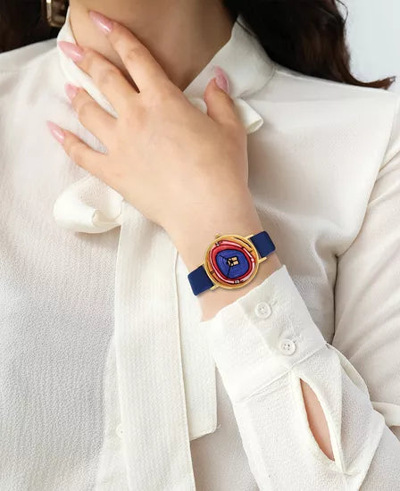 Eine Person trägt eine weiße Bluse und die Friedensreich Hundertwasser Künstler-Armbanduhr „Zeitfinder Seelenbaum“, deren farbenfrohes künstlerisches Gesicht sichtbar ist, wenn ihre Hand nahe ihrem Hals ruht.