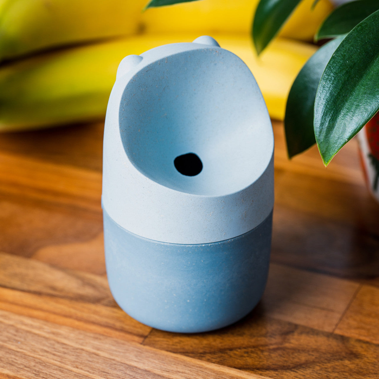 Der Fruchtfliegenfänger KONNA - Blau, eine kleine hellblaue Tasse mit Bärenmotiv, steht auf einer Holzfläche neben Bananen und einer Pflanze.