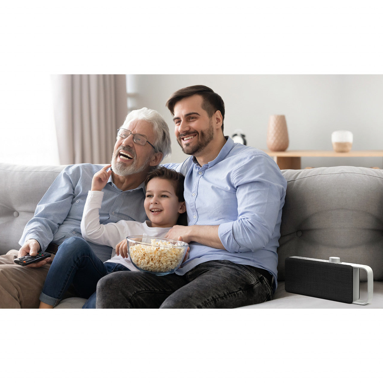Drei Generationen einer Familie lachen und sehen gemeinsam auf der Couch fern und genießen dabei klaren Klang mit dem tragbaren TV-Sprachverstärker von OSKAR.