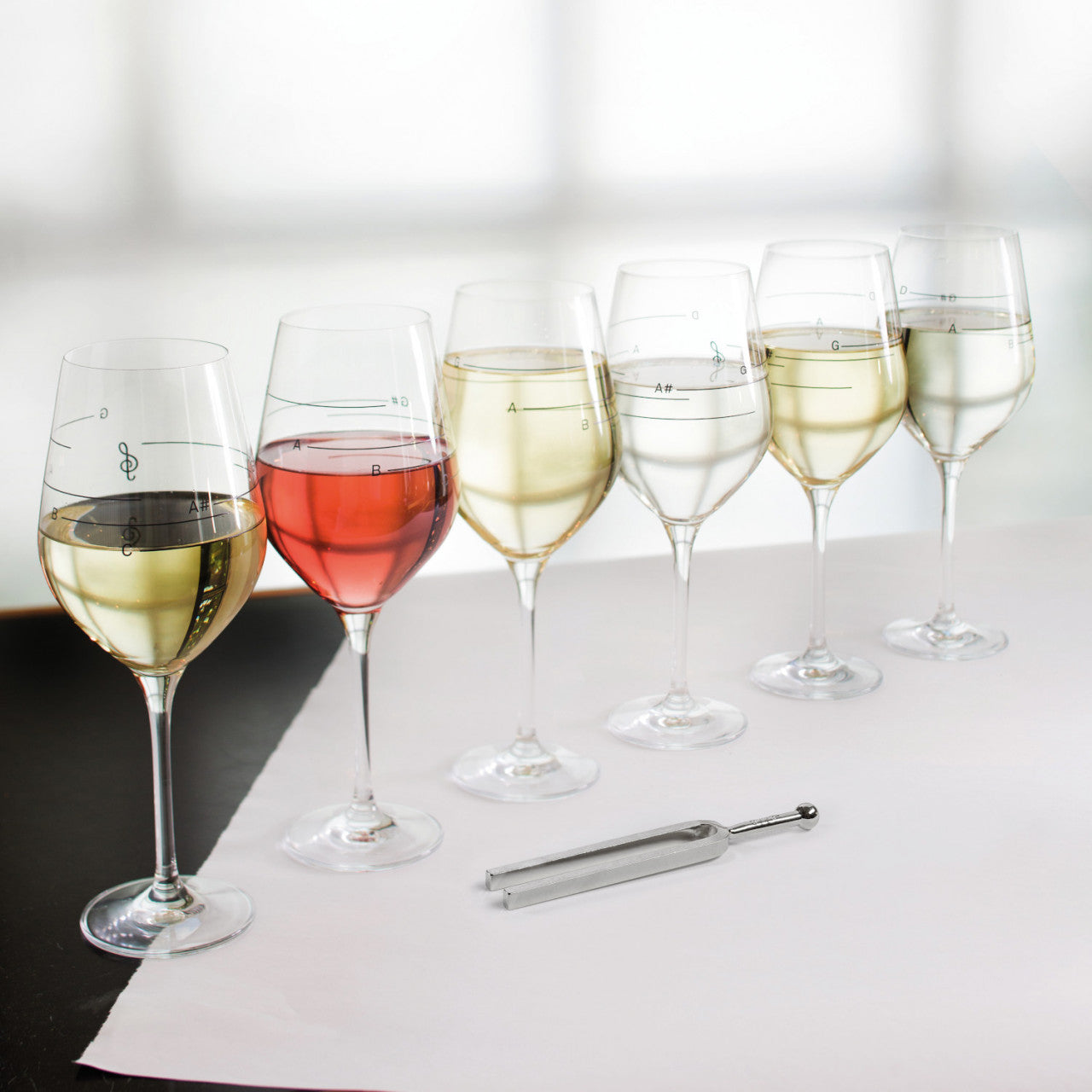 Sechs Gläser „Weinglas mit musikalischen Tönen“, unterschiedlich gefüllt mit Rosé- und Weißwein, davor eine Stimmgabel.