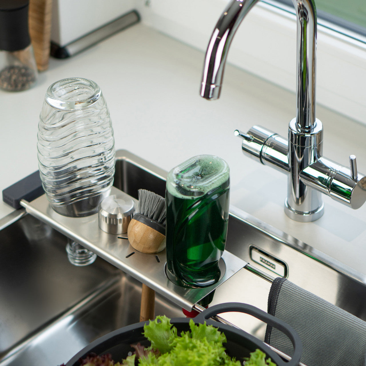 Die Abtropfhilfe Drip.it hält Gläser, eine Bürste und grünes Spülmittel auf einem Metallgestell über frischem Salat in der Küchenspüle.
