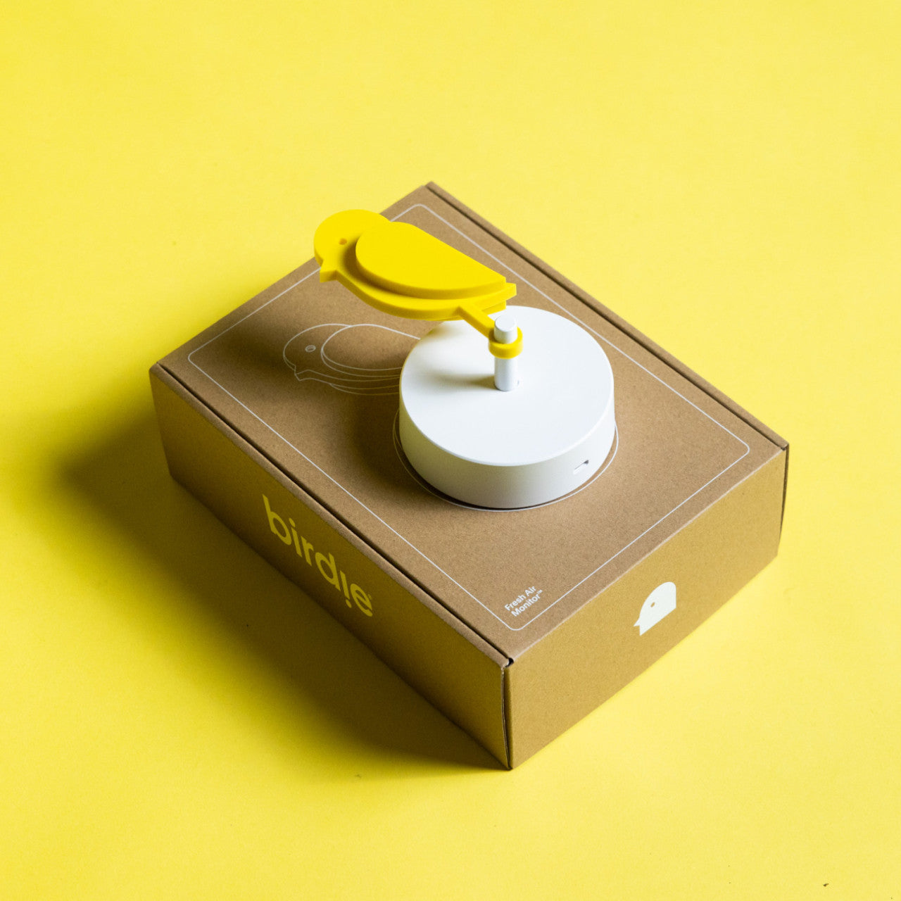 Ein gelber Frischluftmonitor Birdie 2.0 in Vogelform mit CO2-Sensor sitzt auf einem runden weißen Sockel auf einer braunen Box mit der Aufschrift „Birdle“ auf gelbem Hintergrund.