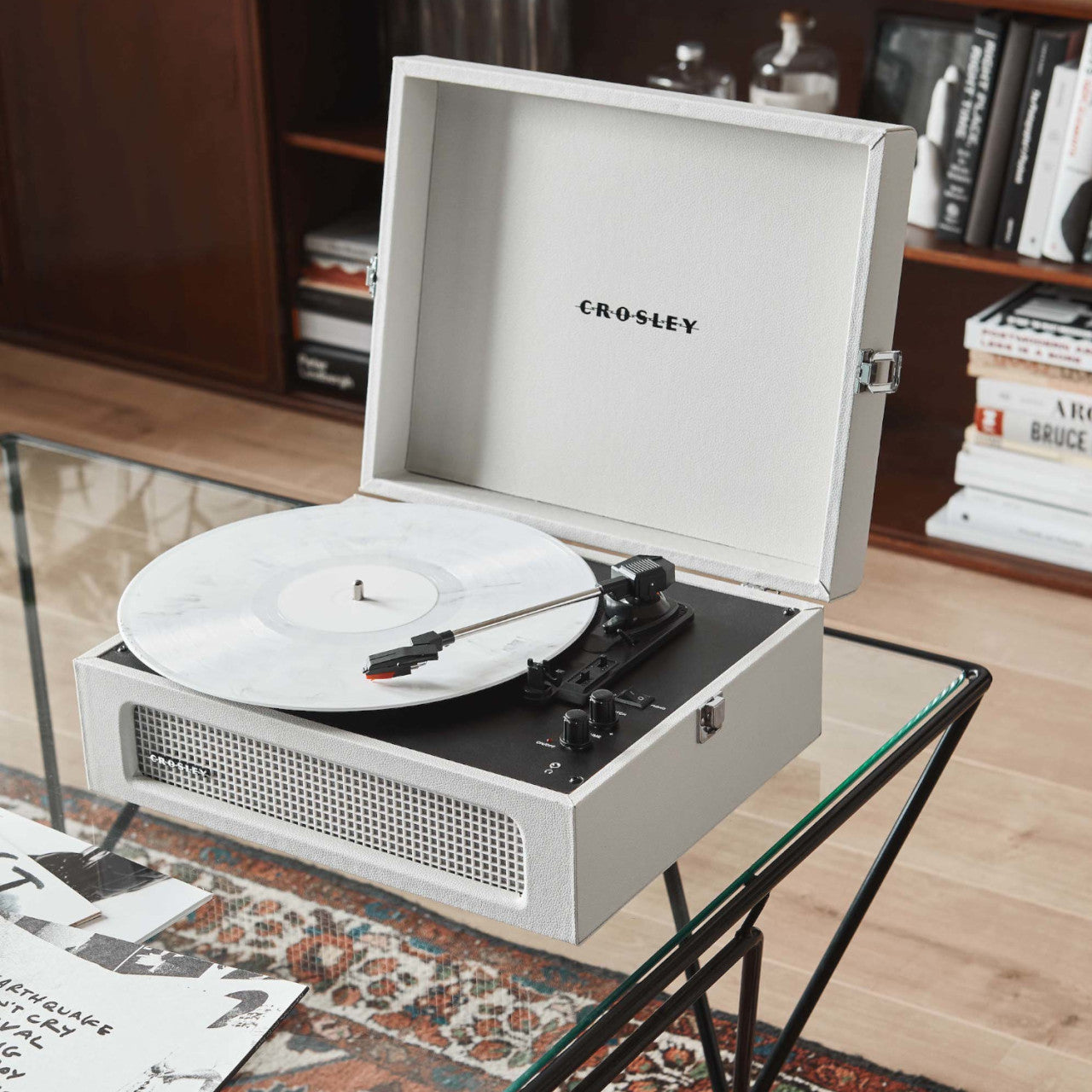 Sandgrauer Crosley Voyager Plattenspieler mit weißem Vinyl auf einem Glastisch im modernen Wohnzimmer-Ambiente.