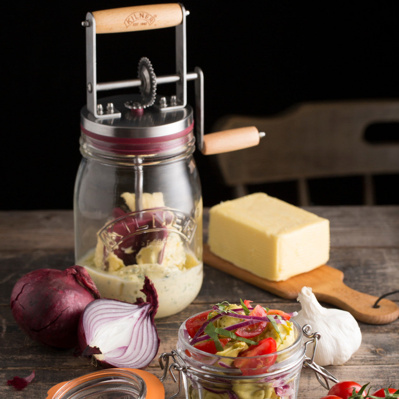 Auf einem Holztisch stehen ein Butterfass (1 Liter), gefüllt mit Butter, ein Stück Butter, rote Zwiebeln, Knoblauch und ein Glas Salat.