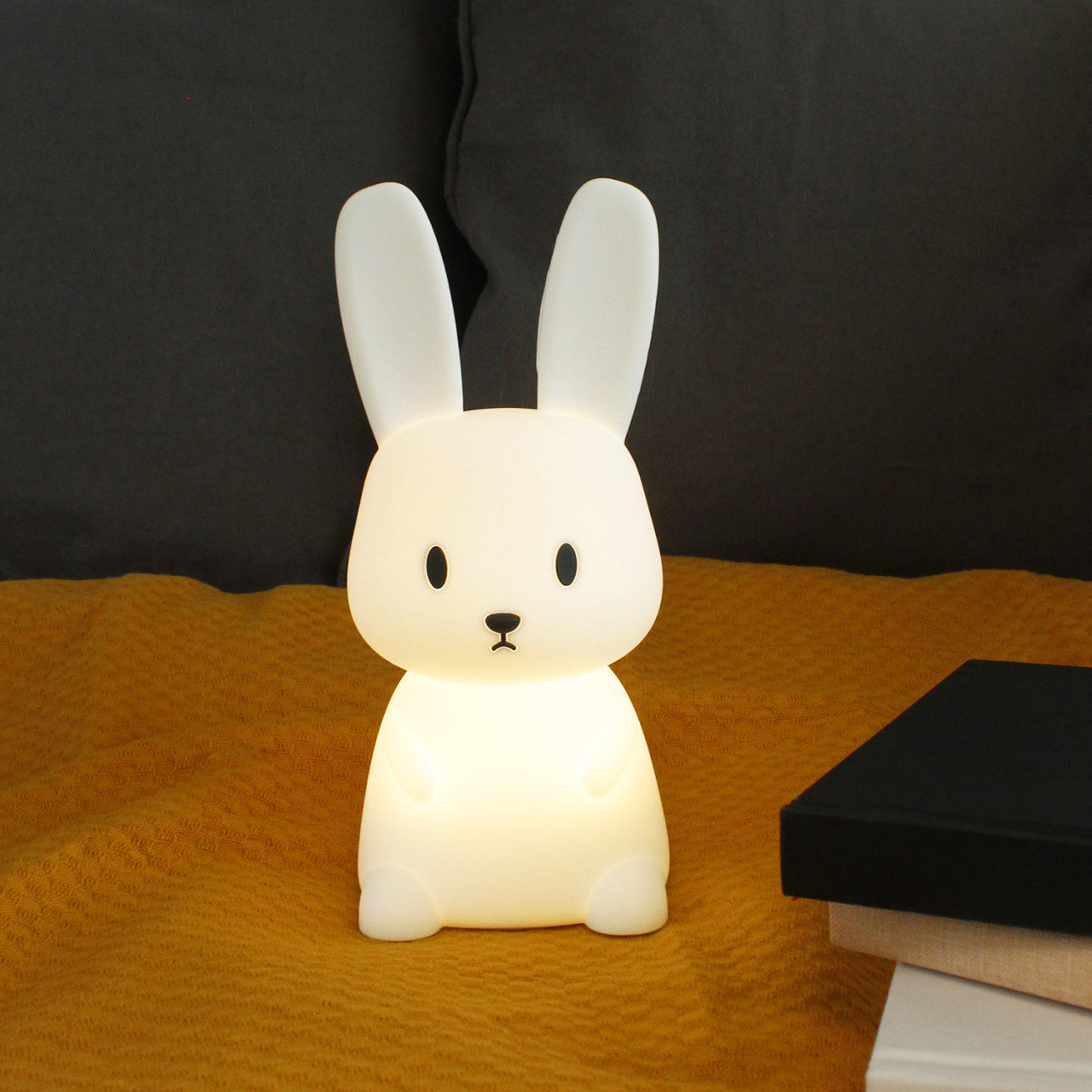 Das Nachtlicht Hase, ein hasenförmiges Nachtlicht, leuchtet sanft auf einer gelben Decke neben einem Stapel Bücher.