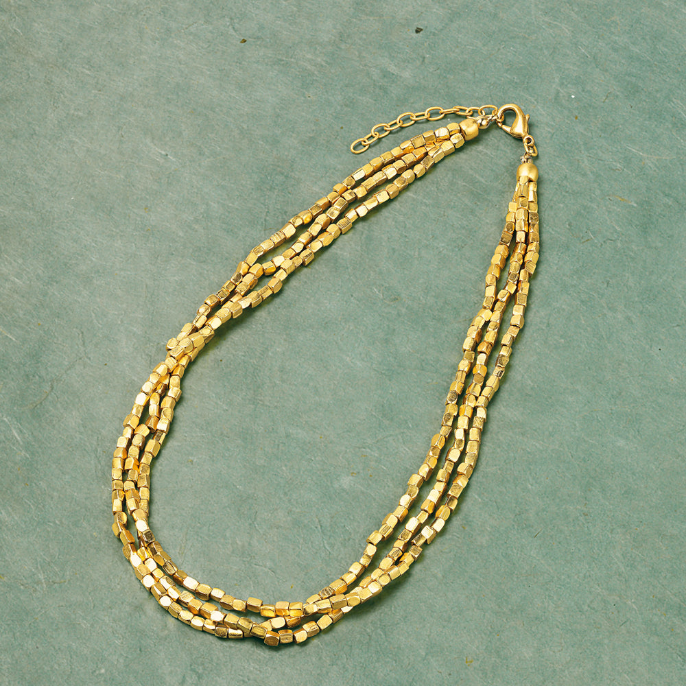 Collier „Golden Nuggets“, dreireihige Halskette aus Goldperlen mit Karabinerverschluss, abgebildet auf strukturiertem grünem Hintergrund.