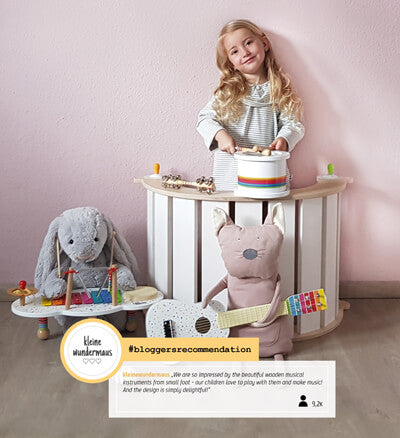 Ein lächelndes Mädchen spielt mit dem hölzernen Musikinstrument Xylophon „Sound“ neben Plüschtieren in einem zartrosa-weißen Raum.