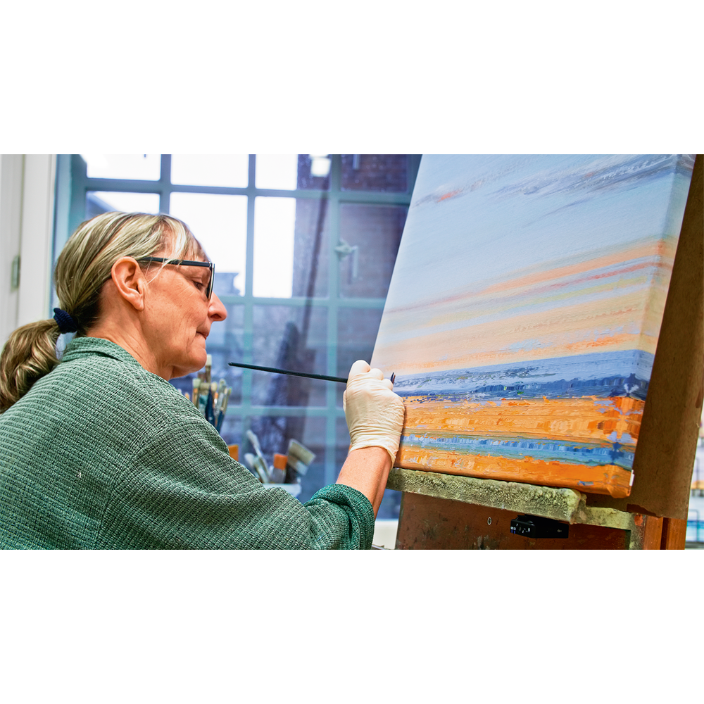 Eine Frau mit Brille malt in einem gut beleuchteten Kunstatelier Anja Strucks „Die Liebe zum Meer“ (2023), eine farbenfrohe Landschaft, auf Leinwand.