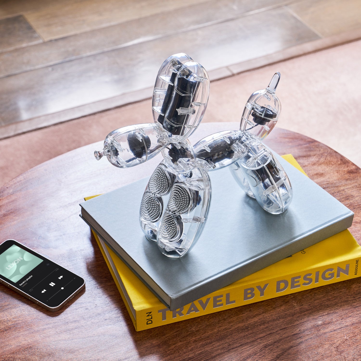 Ein durchsichtiger Lautsprecher in Form eines Ballonhundes liegt auf einem Holztisch auf Büchern neben einem Smartphone.