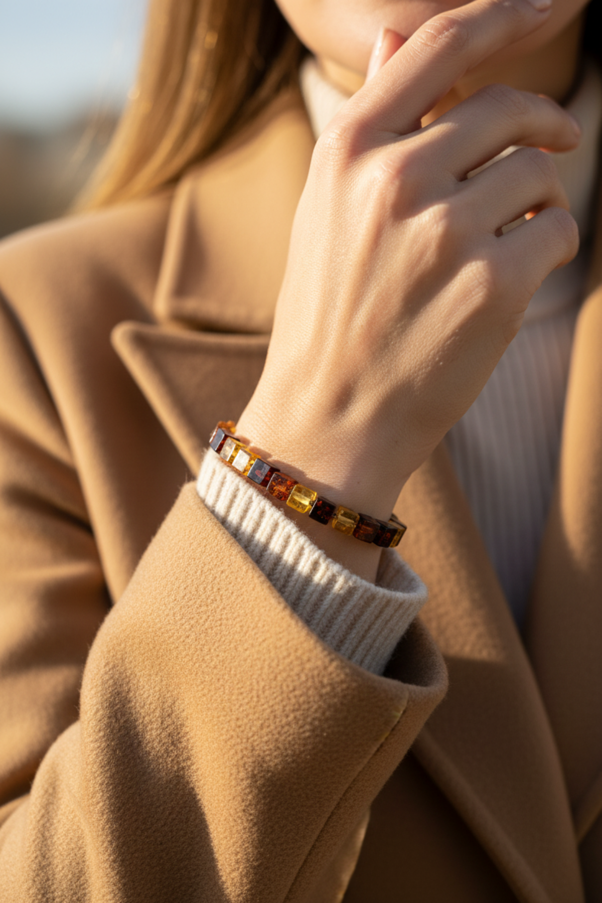 Eine Person, die einen hellbraunen Mantel und einen gerippten Pullover trägt, ist mit einem quadratischen Perlenarmband am Handgelenk abgebildet und hält ihre Hand nahe am Gesicht.