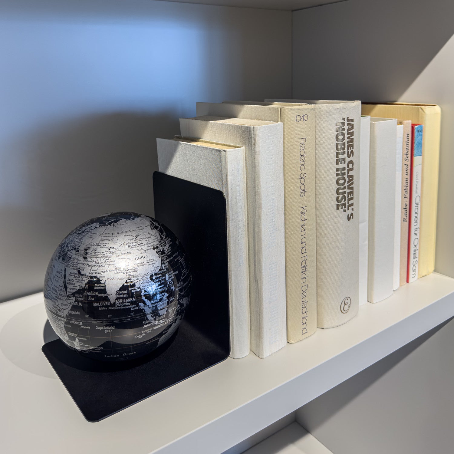 Eine schwarz-silberne Globus-Buchstütze stützt eine Reihe weißer Bücher in einem weißen Bücherregal.