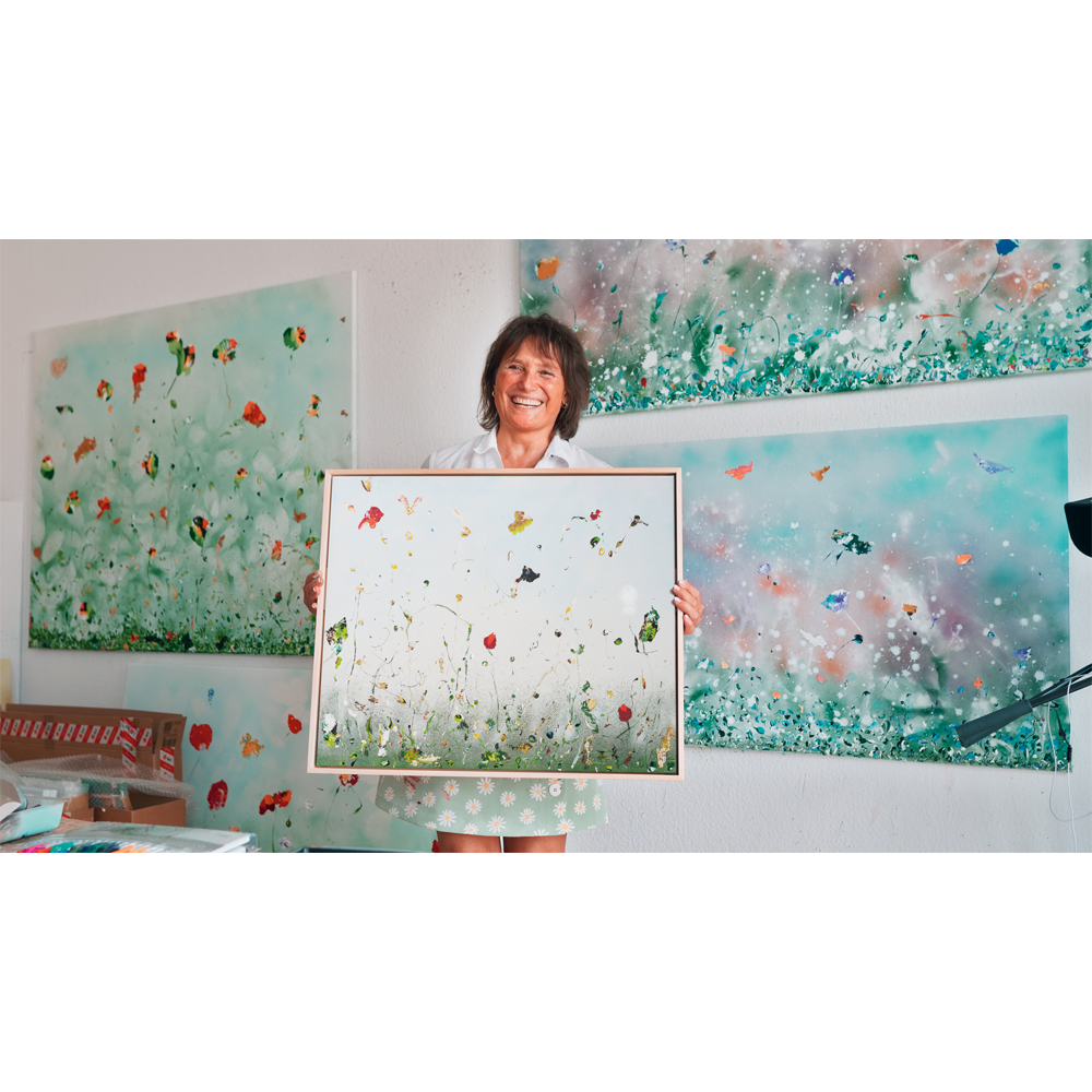 Lächelnde Frau hält „Hommage à Debussy (Bijan) – Jubiläums-Edition“ (2020) von Christine Müller, ein gerahmtes Serienunikat, vor ähnlichen Kunstwerken.
