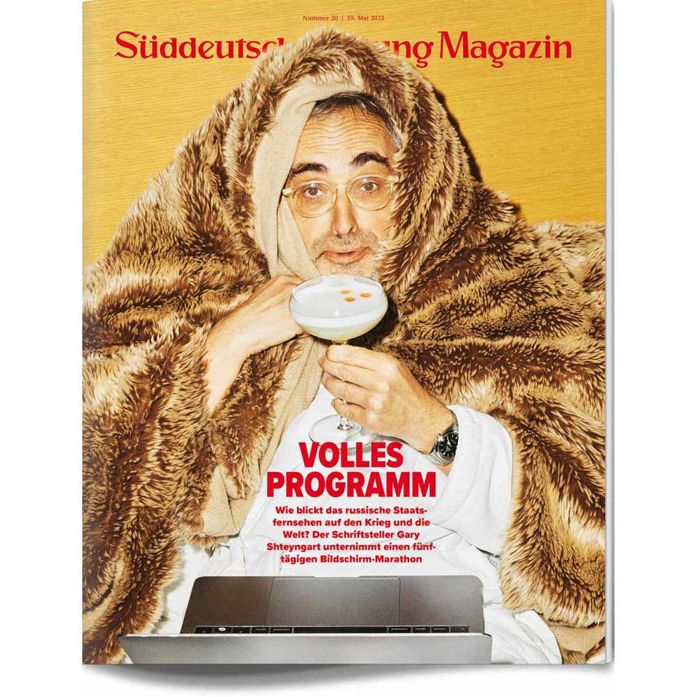 Ein in eine Decke gehüllter Mann hält einen Cocktail in der Hand und sitzt hinter einem Laptop auf einem Zeitschriftencover.