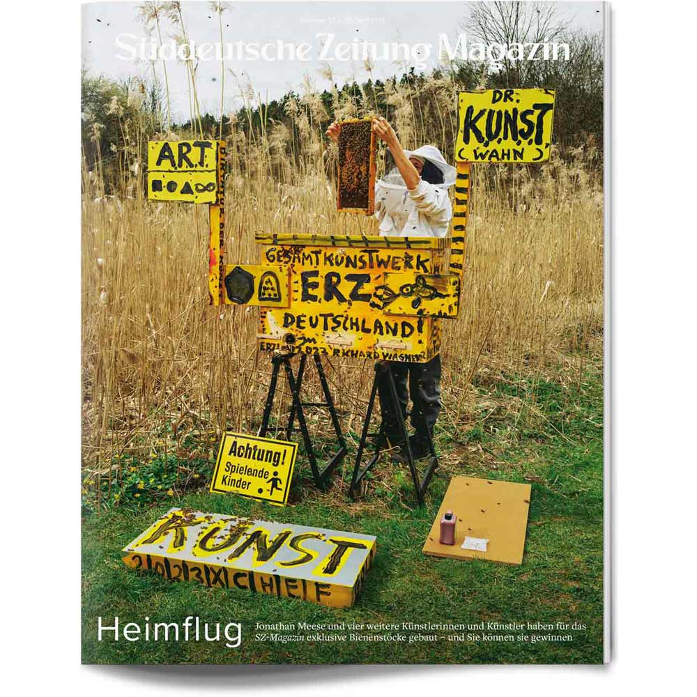 Eine Person in der Natur, die gelbe Schilder mit deutschen Worten malt, umgeben von hohem Gras und handgemachten Kunstwerken.