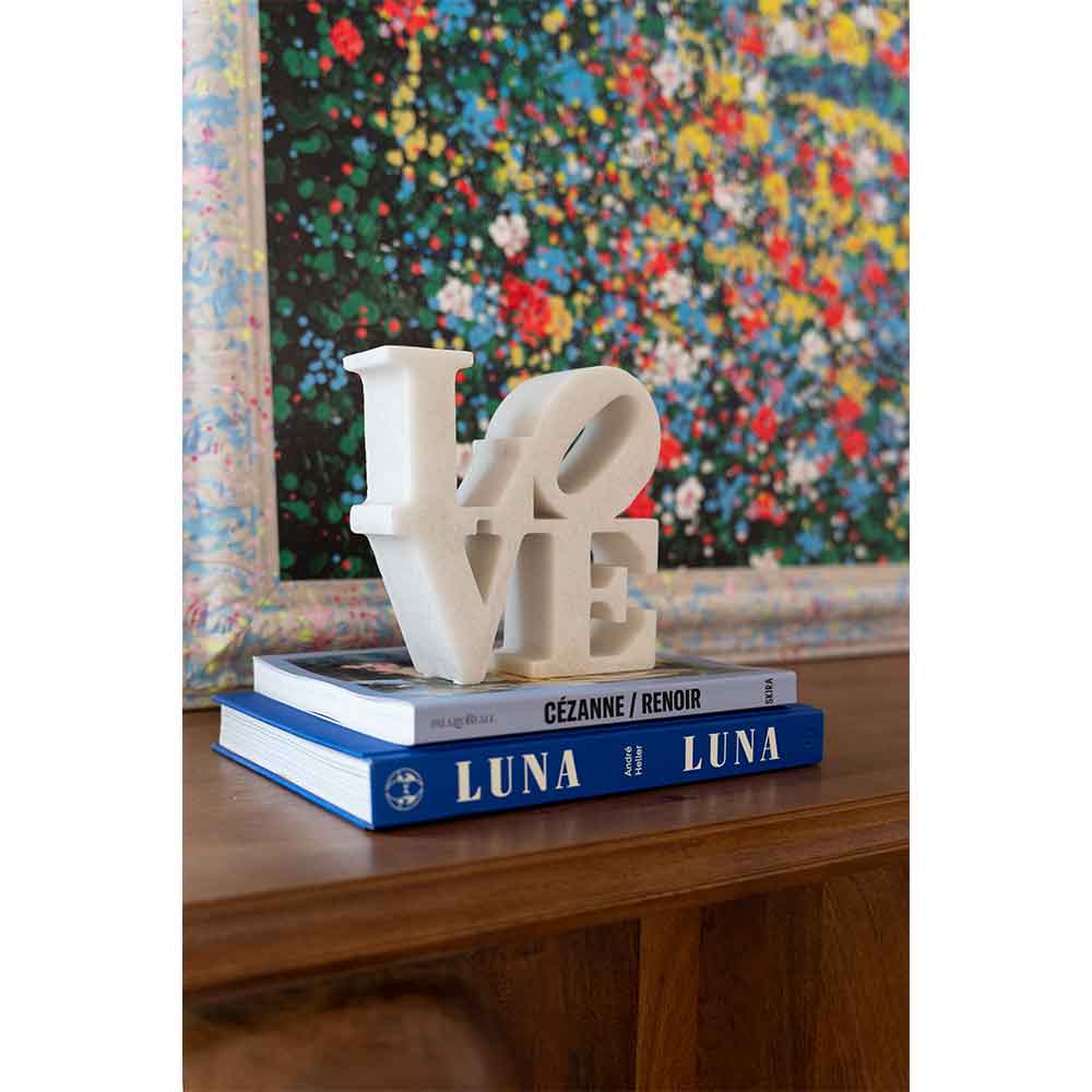Eine weiße LOVE-Skulptur sitzt auf zwei blauen LUNA-Büchern vor einem bunt gerahmten Gemälde.