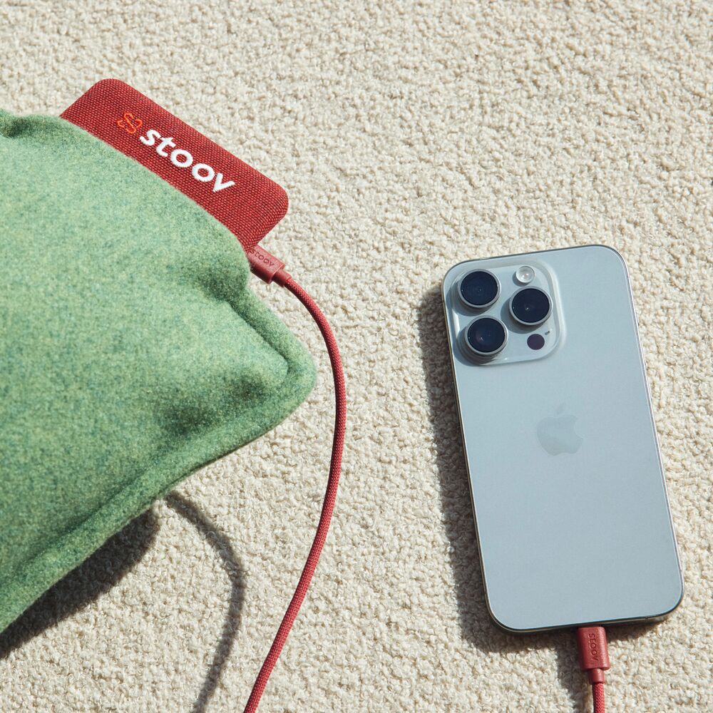 Ein Stoov-Heizkissen und ein silbernes iPhone mit roten Ladekabeln auf einem beigen Teppich.