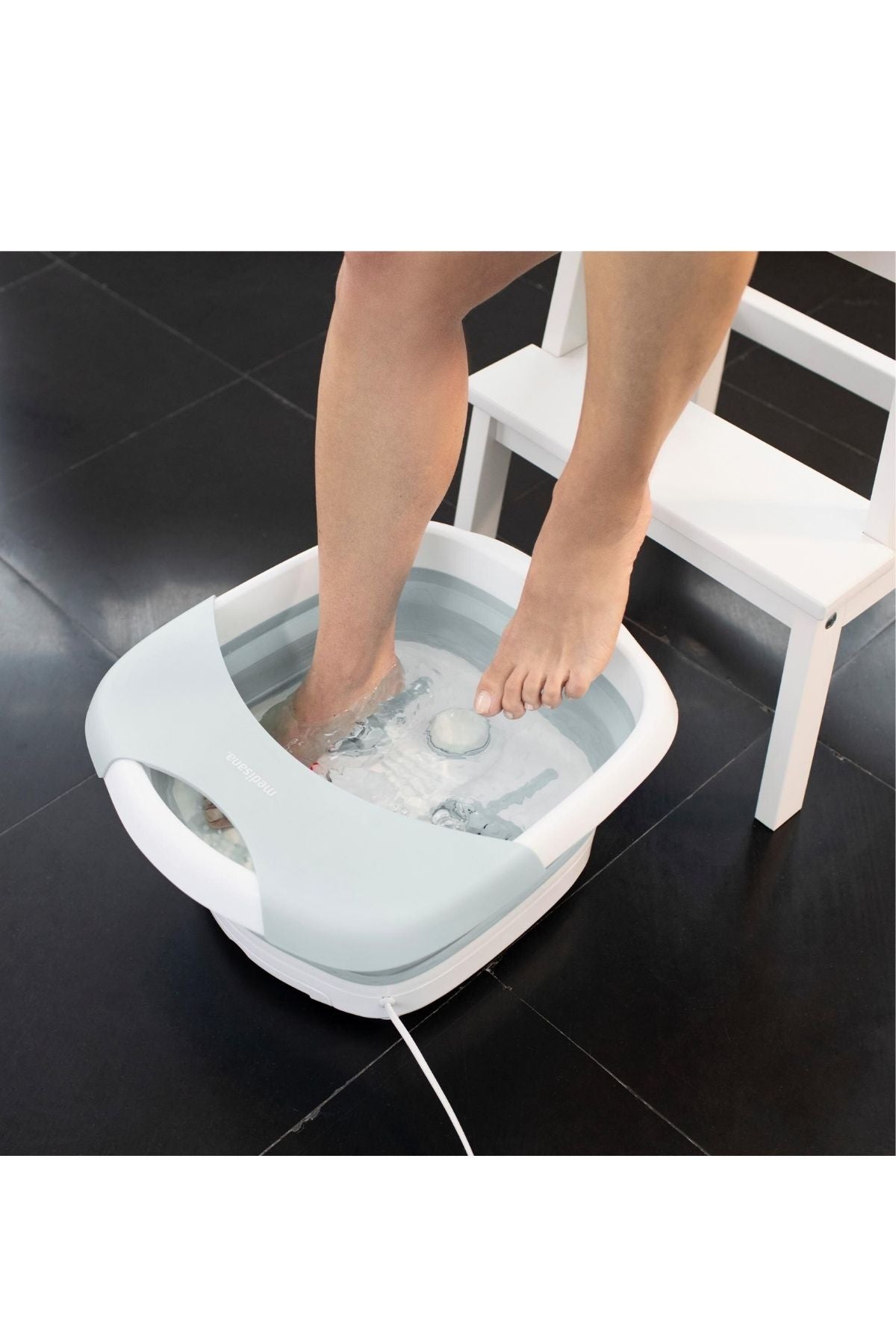 Eine Person legt ihre Füße in ein weißes und hellblaues, mit Wasser gefülltes elektrisches Fußsprudelbad, das auf einem schwarzen Kachelboden neben einem weißen Stuhl steht.