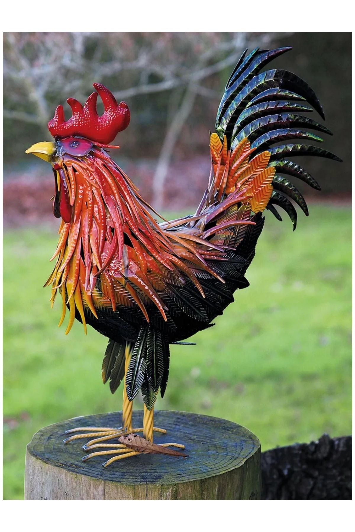 Eine bunte Metallhahnskulptur steht auf einem Baumstumpf im Freien, mit grünem Gras und verschwommenen Bäumen im Hintergrund.
