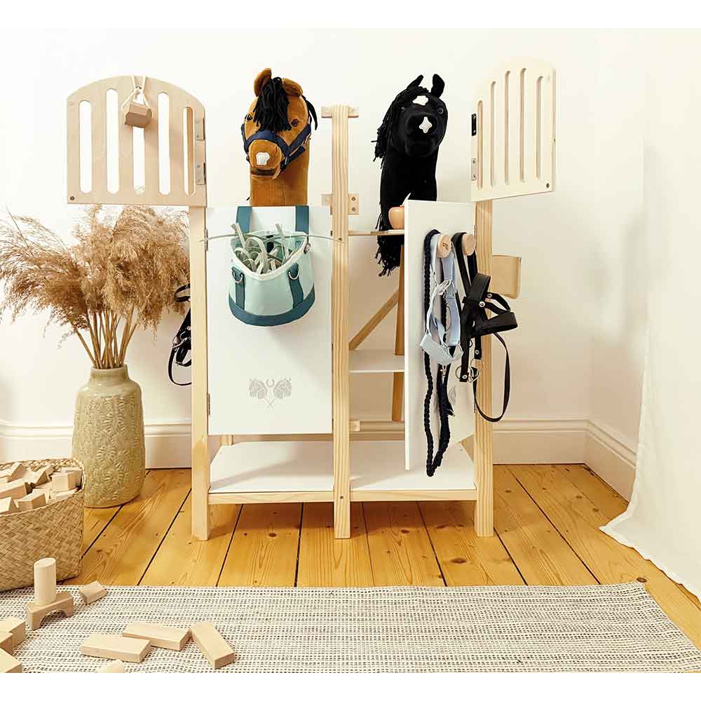 Hobby-Horse-Pferdestall für Kinder mit zwei Spielzeugpferdeköpfen, Zügeln, einem Korb mit Holzklötzen und einer dekorativen Topfpflanze auf einem Holzboden.