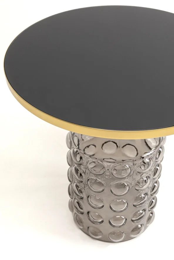 Runder Tisch mit schwarzer, glänzender Platte, goldener Kante und einem strukturierten Glasfuß mit Blasenmuster.