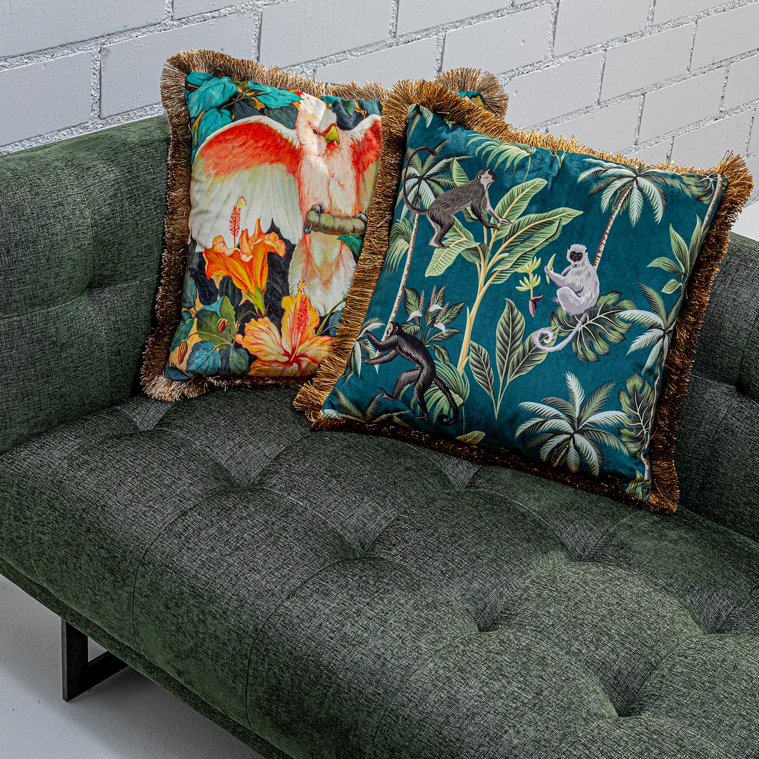 Zwei farbenfrohe dekorative Kissen mit Tier- und Pflanzenmotiven auf einem grünen getufteten Sofa vor einer weißen Backsteinwand.